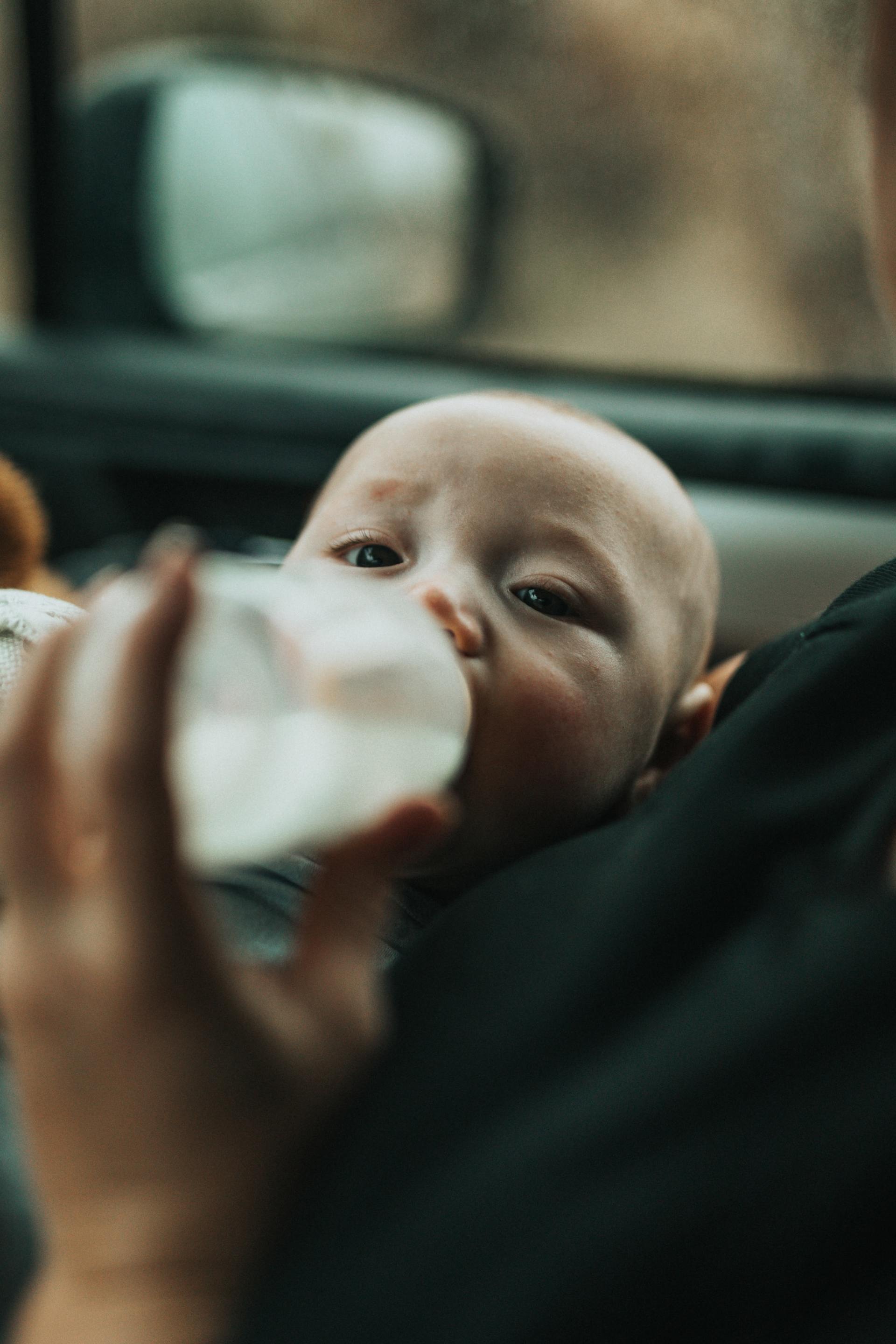 Baby drinking from a bottle, held by a person in a vehicle.