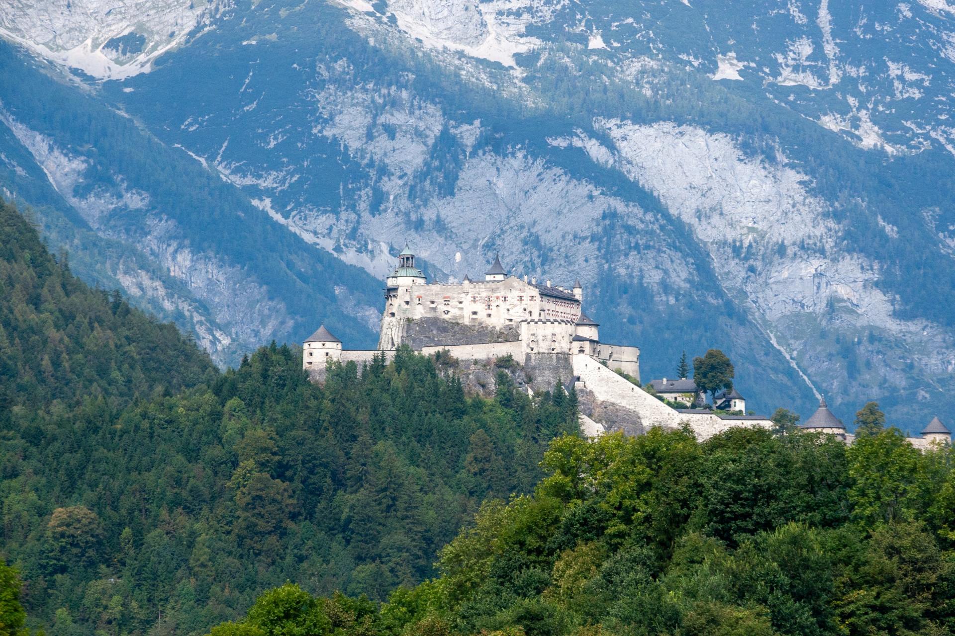 Weiße Festung an einem bewaldeten Berghang mit schneebedeckten Gipfeln im Hintergrund