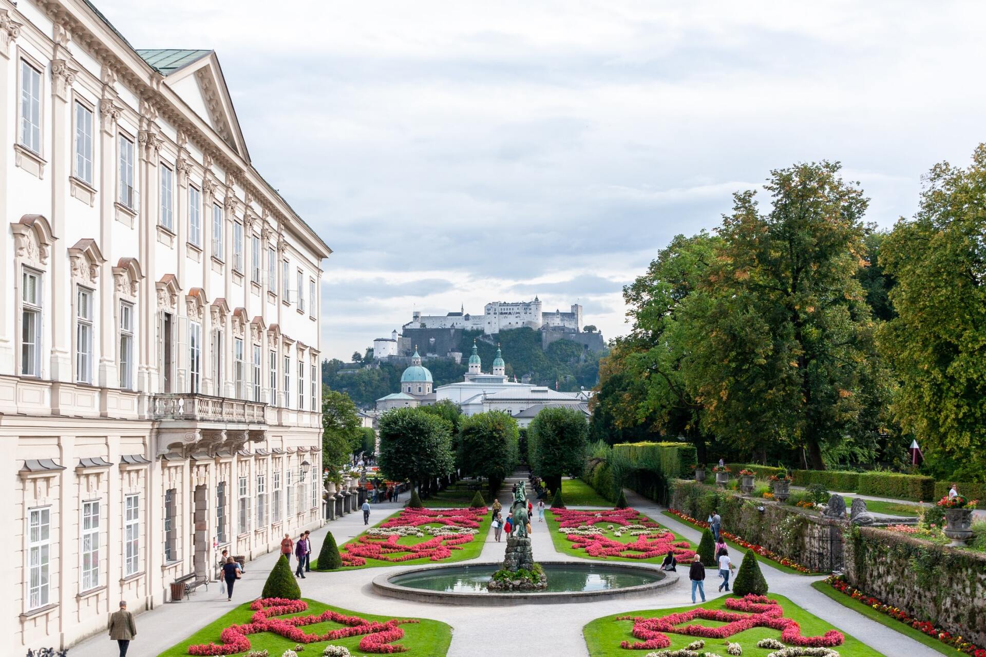 Ein formaler Garten mit rosa Blumenbeeten, links ein weißes Schloss und auf dem fernen Hügel eine Burg.