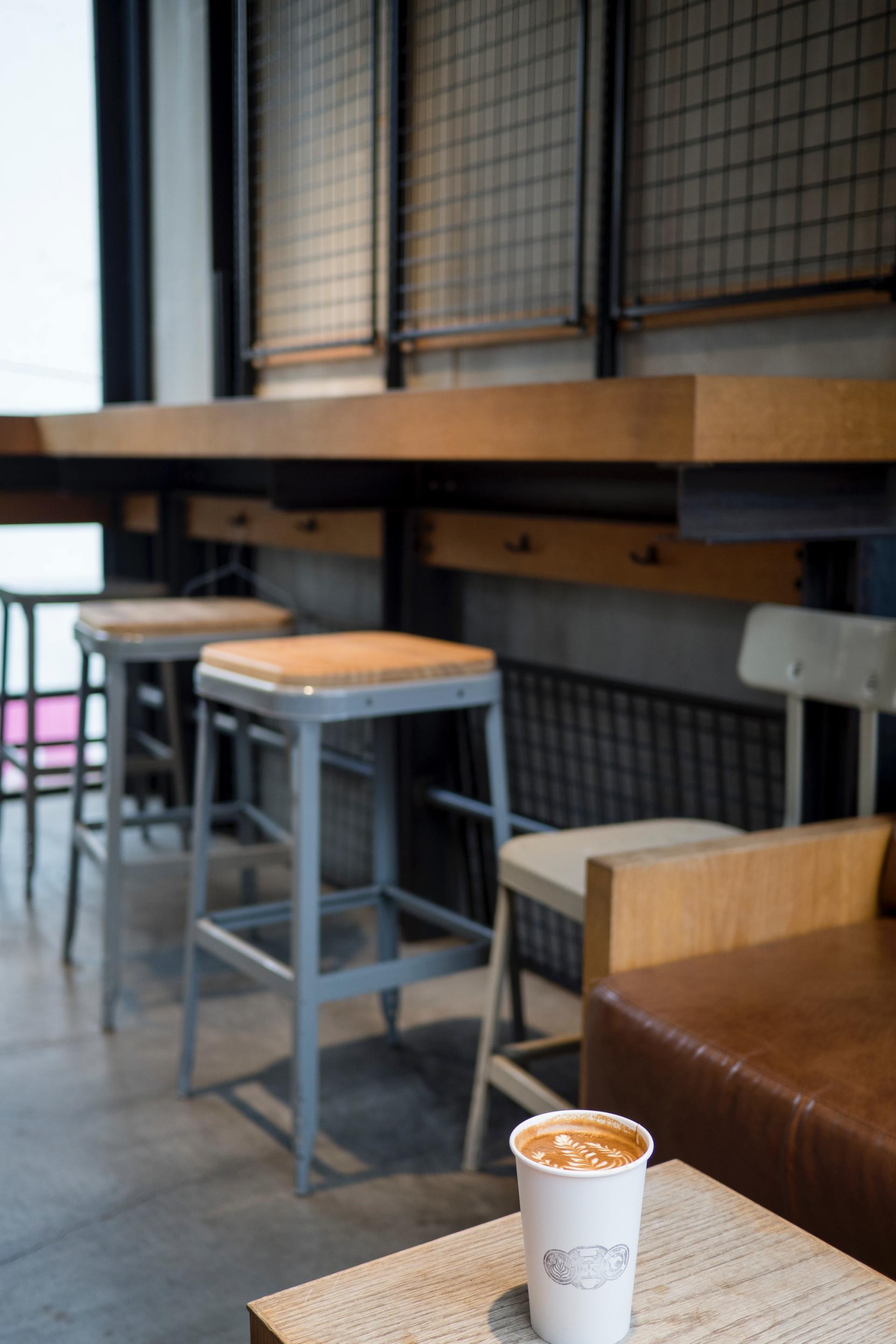 In un caffè moderno in stile industriale, una tazza di carta bianca è appoggiata su un tavolino davanti a degli sgabelli e a un bancone di legno.