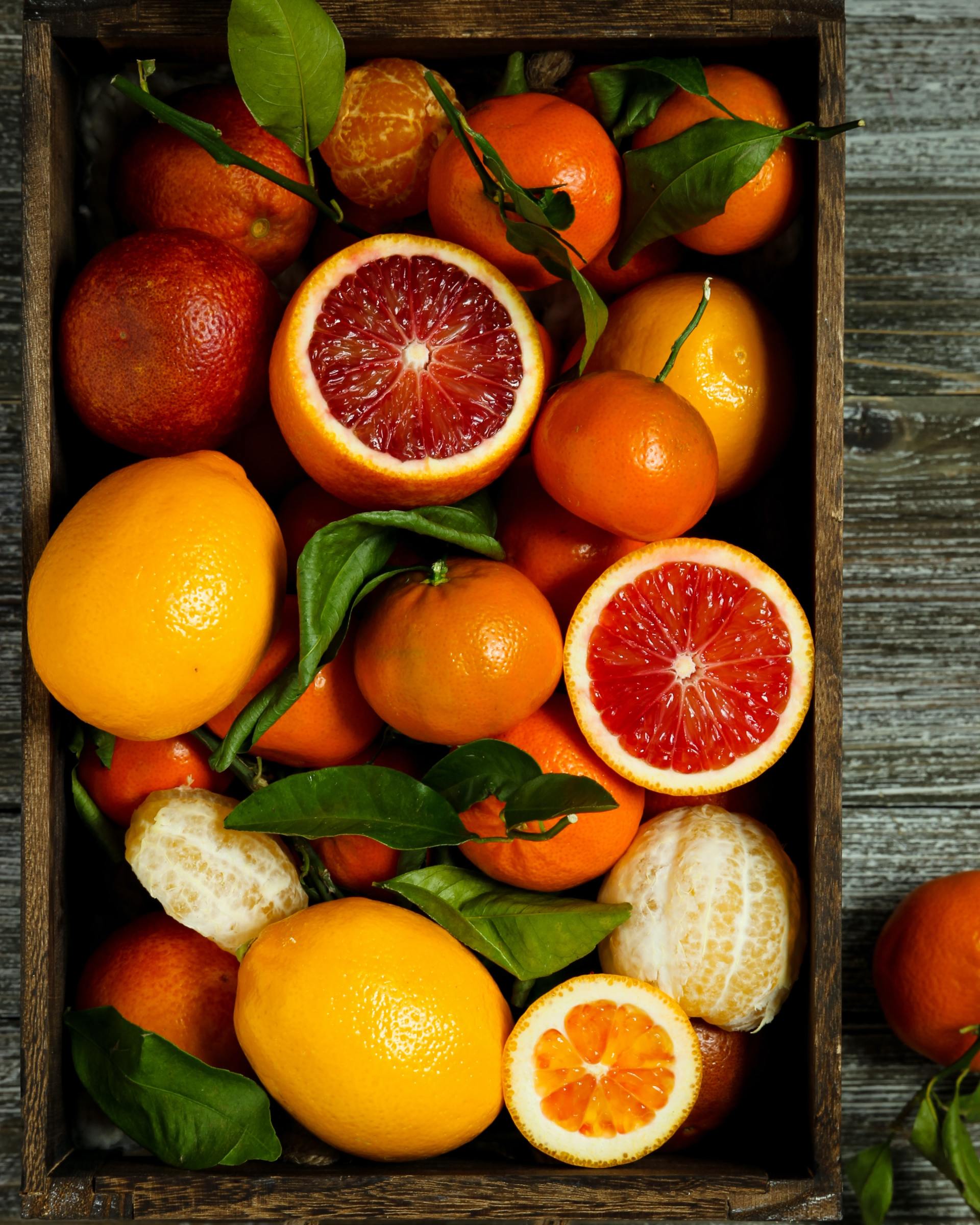 Wooden crate filled with various citrus fruits: oranges, blood oranges, tangerines, and lemons, some with green leaves.