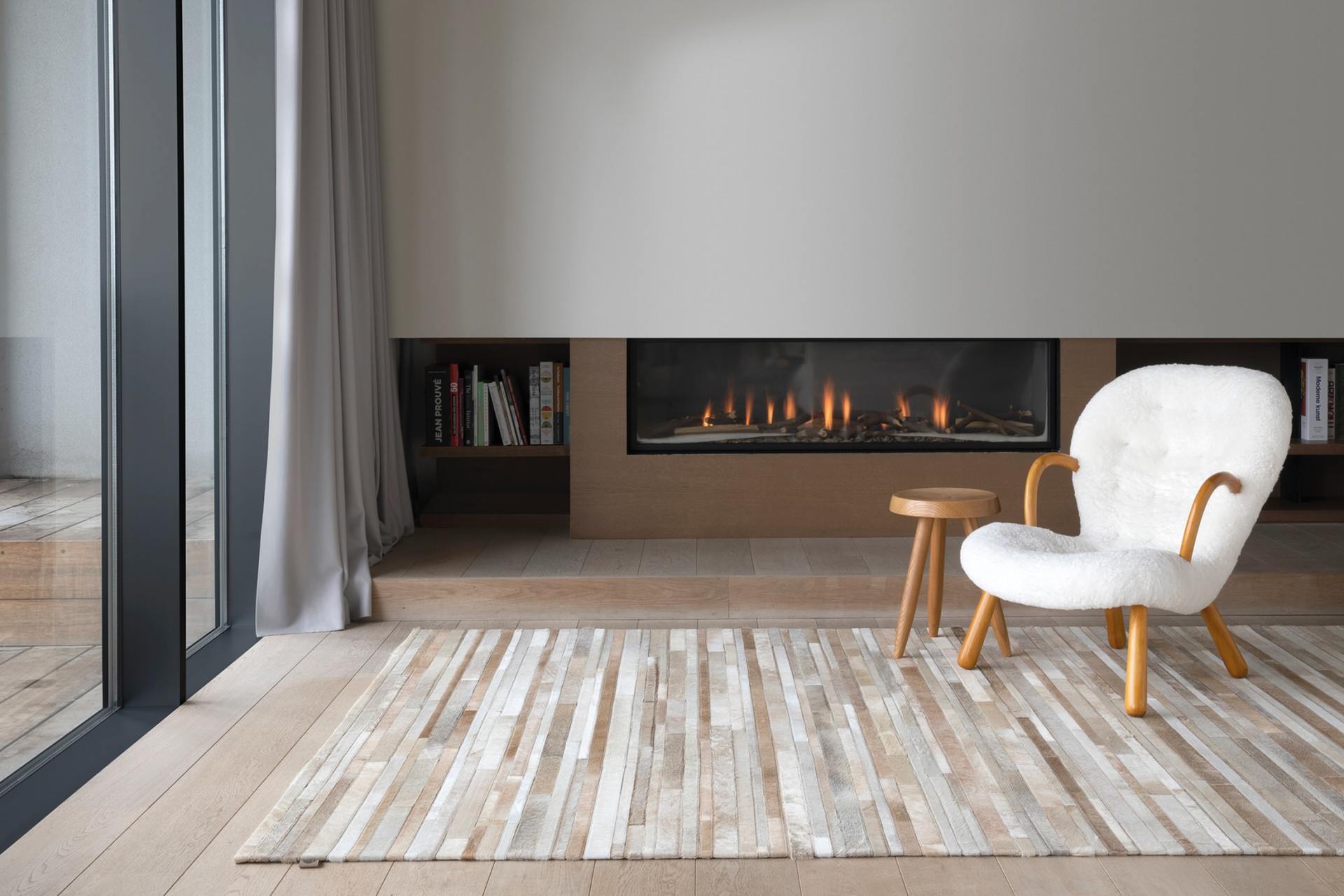 Cozy living room with fireplace, white fluffy chair, wooden stool, and a patterned rug on a hardwood floor.