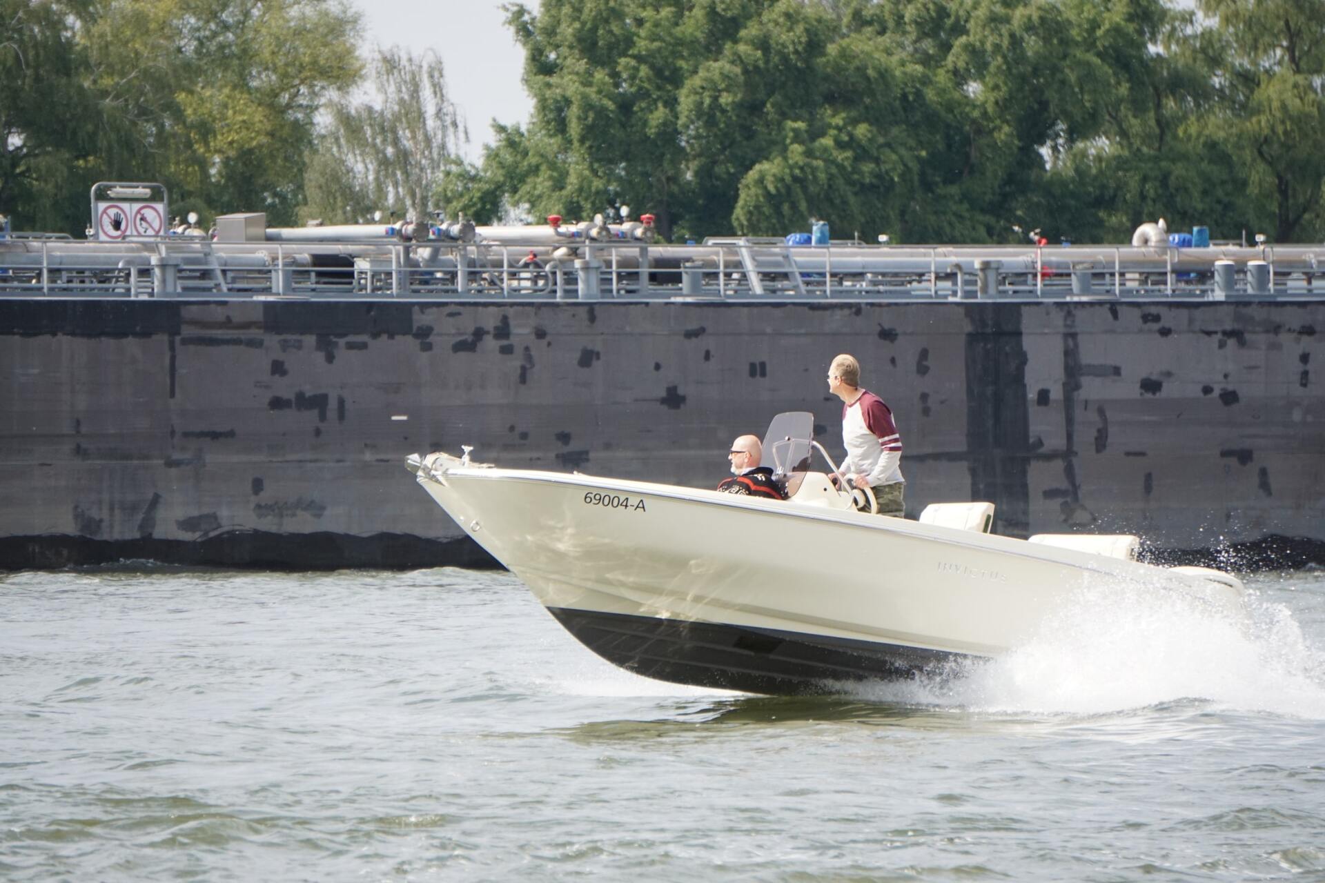 Kleines Motorboot rast mit zwei Personen an Bord über das Wasser neben einer Betonmauer.