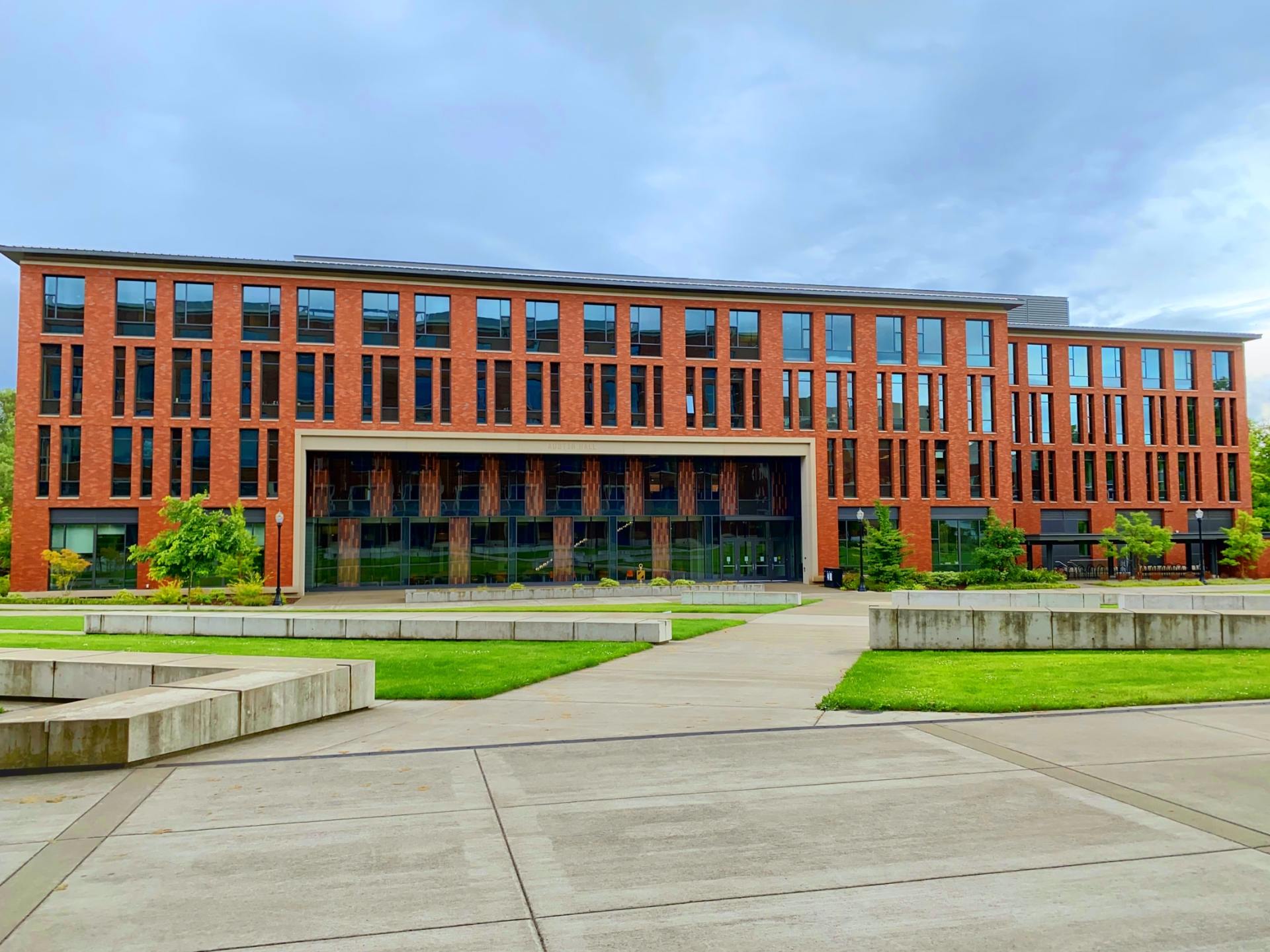 Brick building with rows of windows, glass entrance, and grassy courtyard under a cloudy sky.