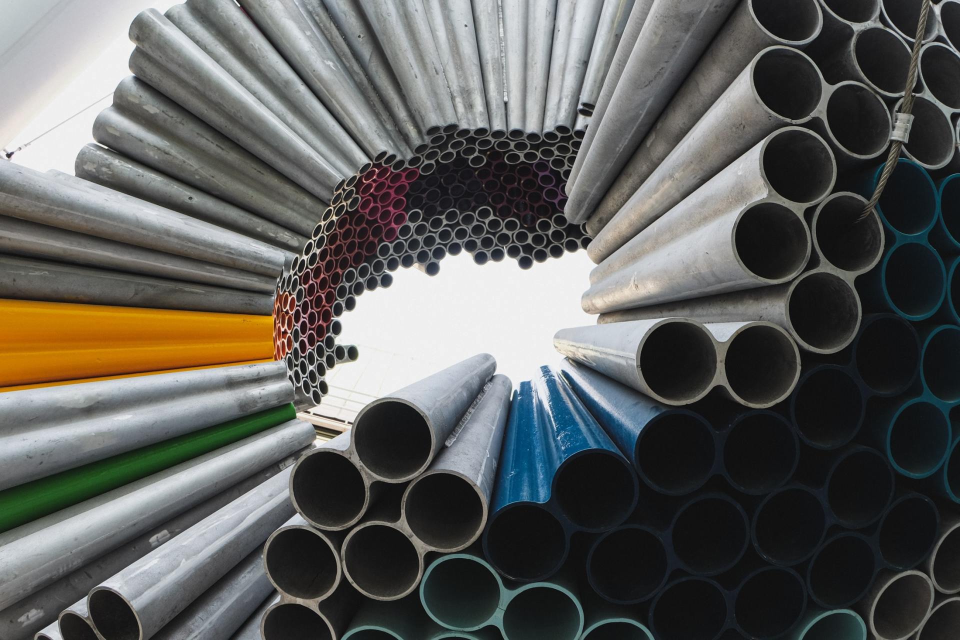 Una disposizione circolare di tubi industriali, di colori e dimensioni diverse, crea uno spazio aperto al centro, stagliandosi contro un cielo chiaro.