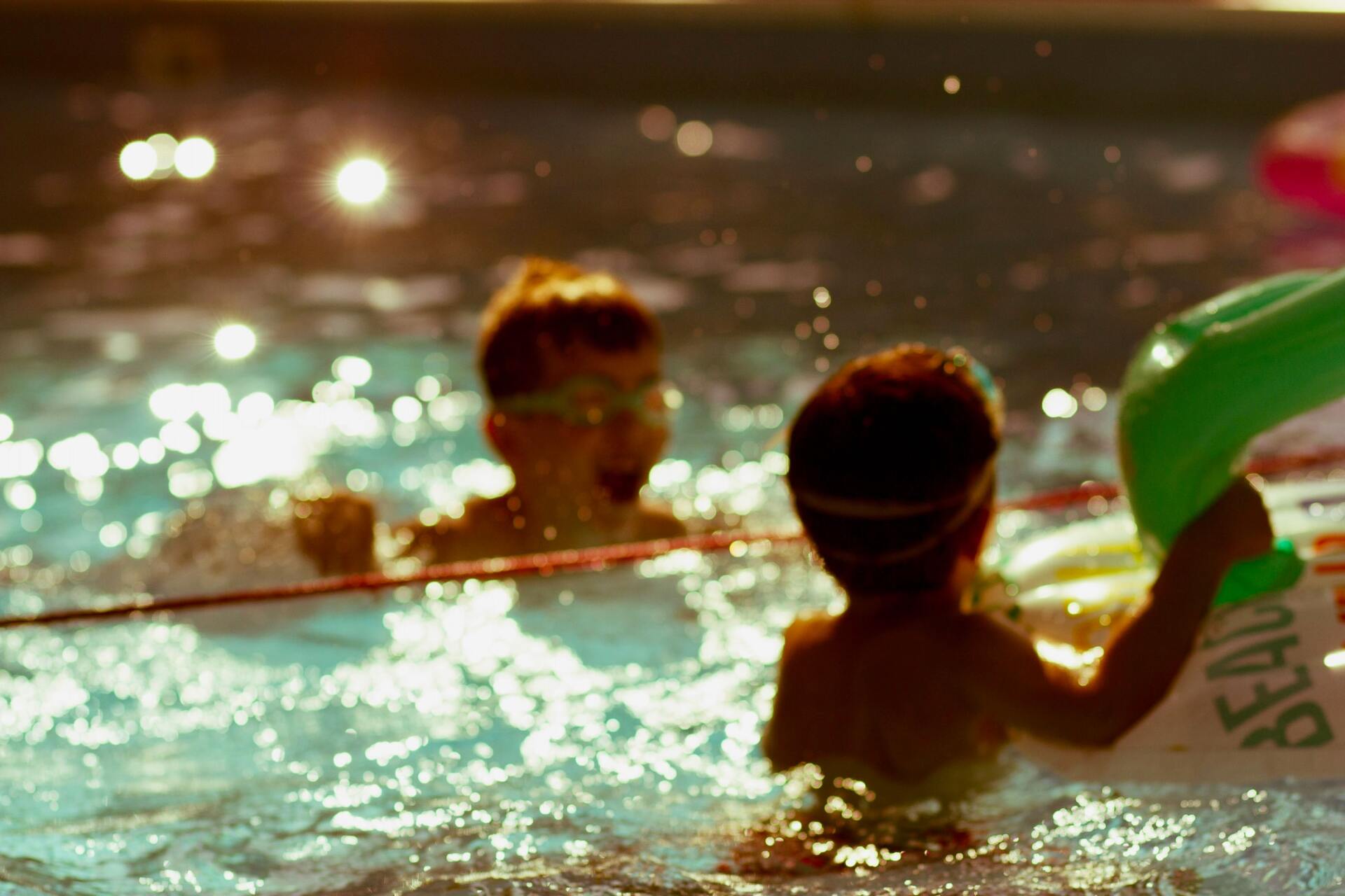 Kinder spielen in der Abenddämmerung in einem glitzernden Schwimmbecken, im Vordergrund eine grüne Schwimminsel.