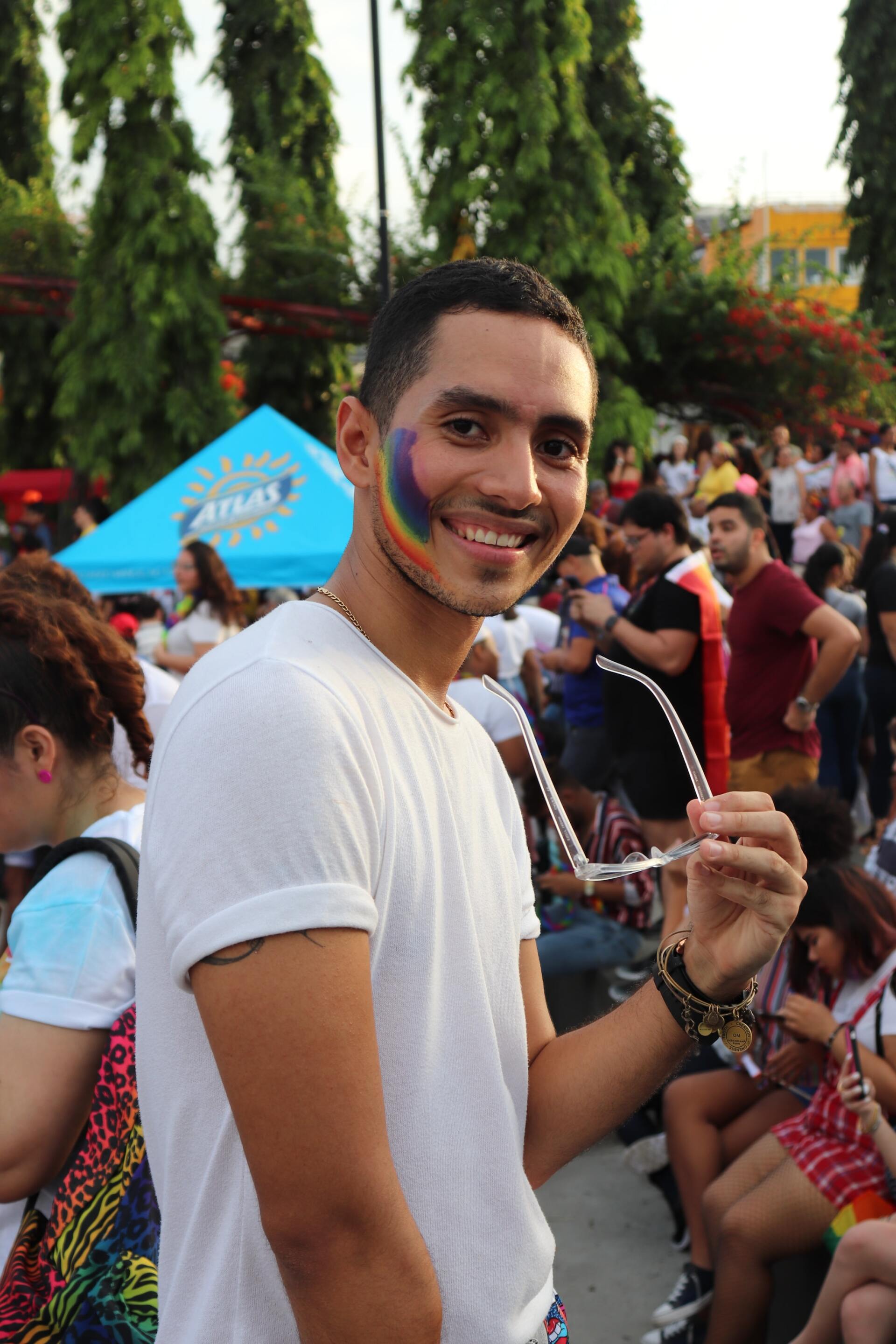 Man with rainbow face paint smiles, holding glasses, at outdoor Pride event.