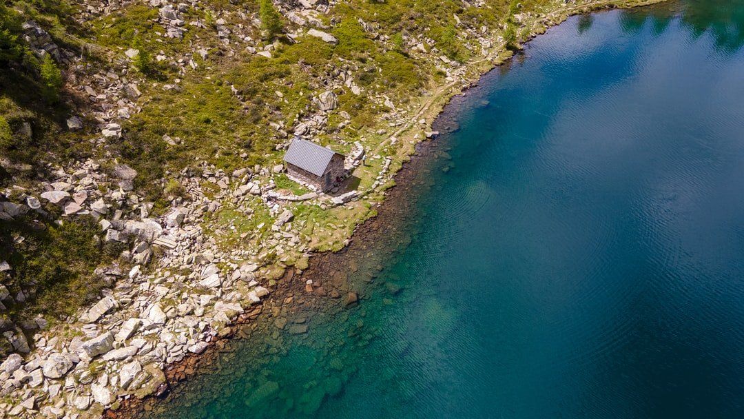 Kleine Hütte an einem felsigen, grünen Ufer neben klarem, blauem Wasser, Luftbild