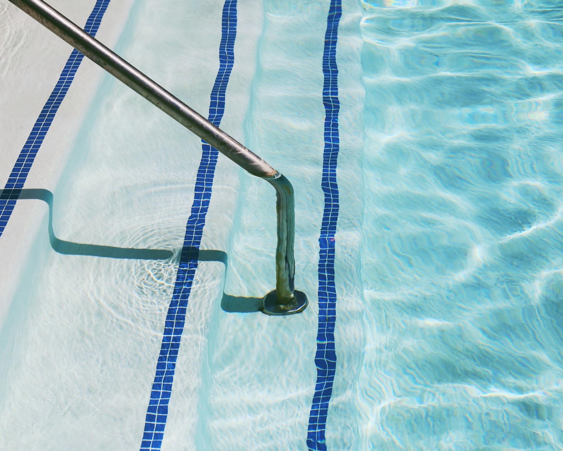 Swimming pool steps submerged in crystal blue water. A silver handrail proceeds down the steps. Clear blue water creates a nice ripple effect.