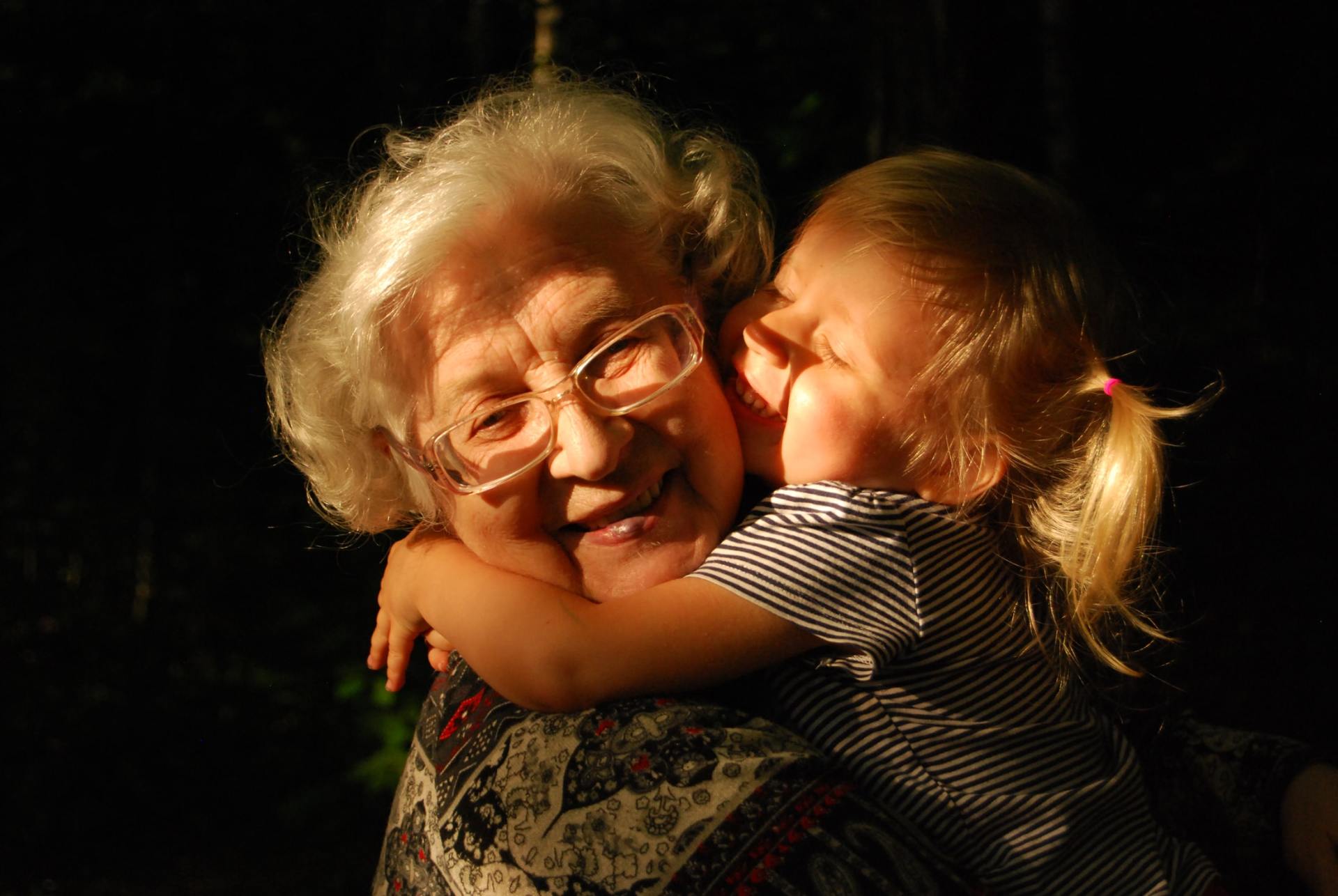 A child hugs and kisses a smiling older person wearing glasses; both are outside in the sunlight.