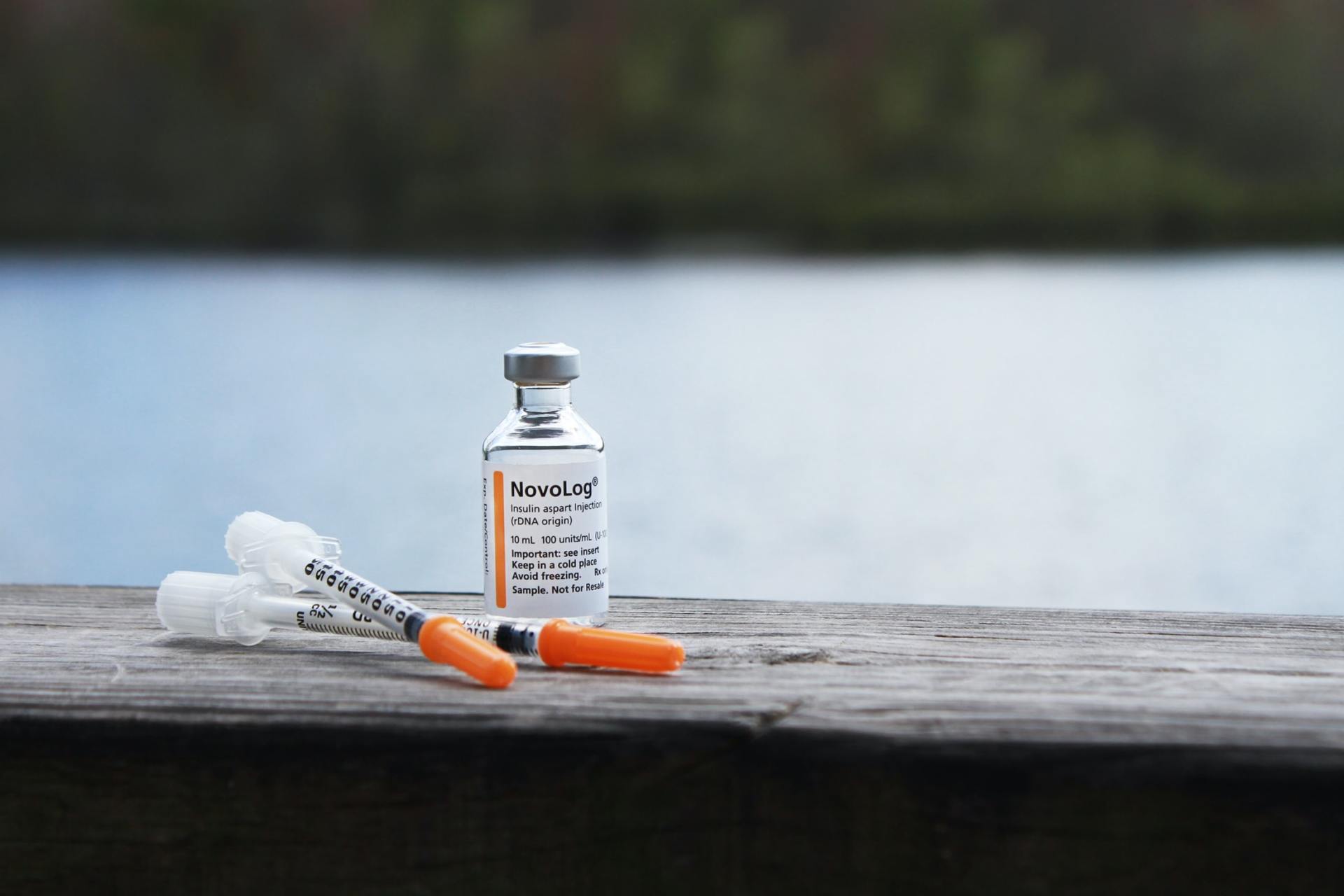 A vial of medicine and two syringes rest on a wooden surface, with a blurred lake and forest in the background.