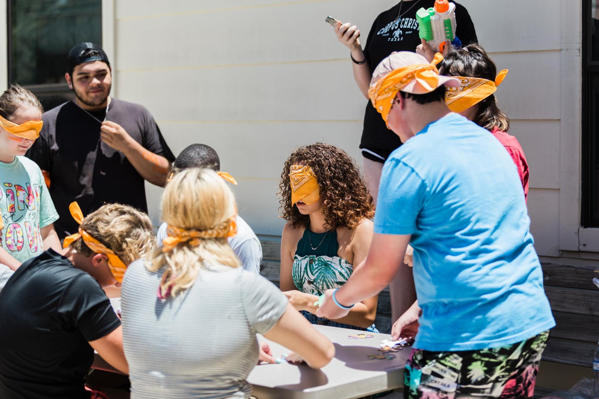 Group of people playing a game outdoors. Many are blindfolded, some are holding cards, and others are observing.