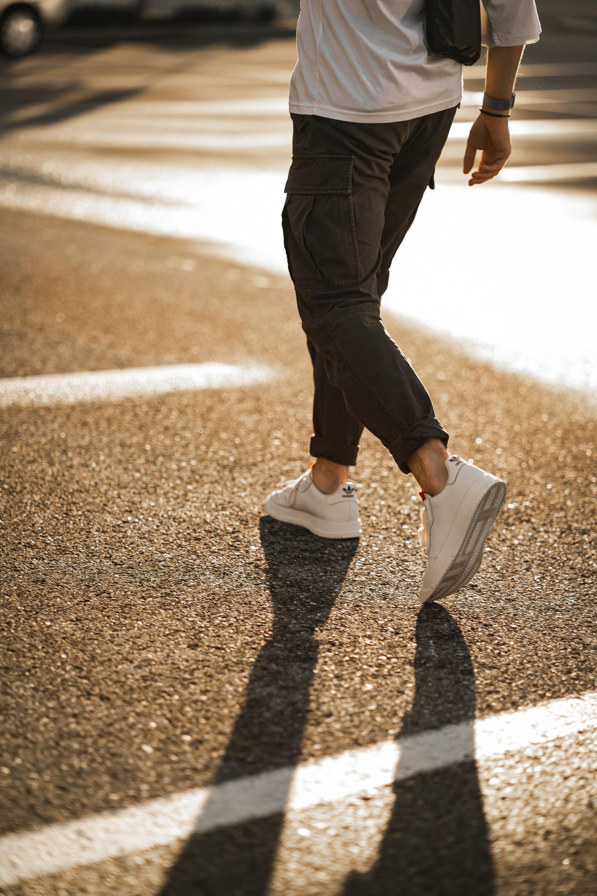 Person walking across a sunlit street in white sneakers and dark pants