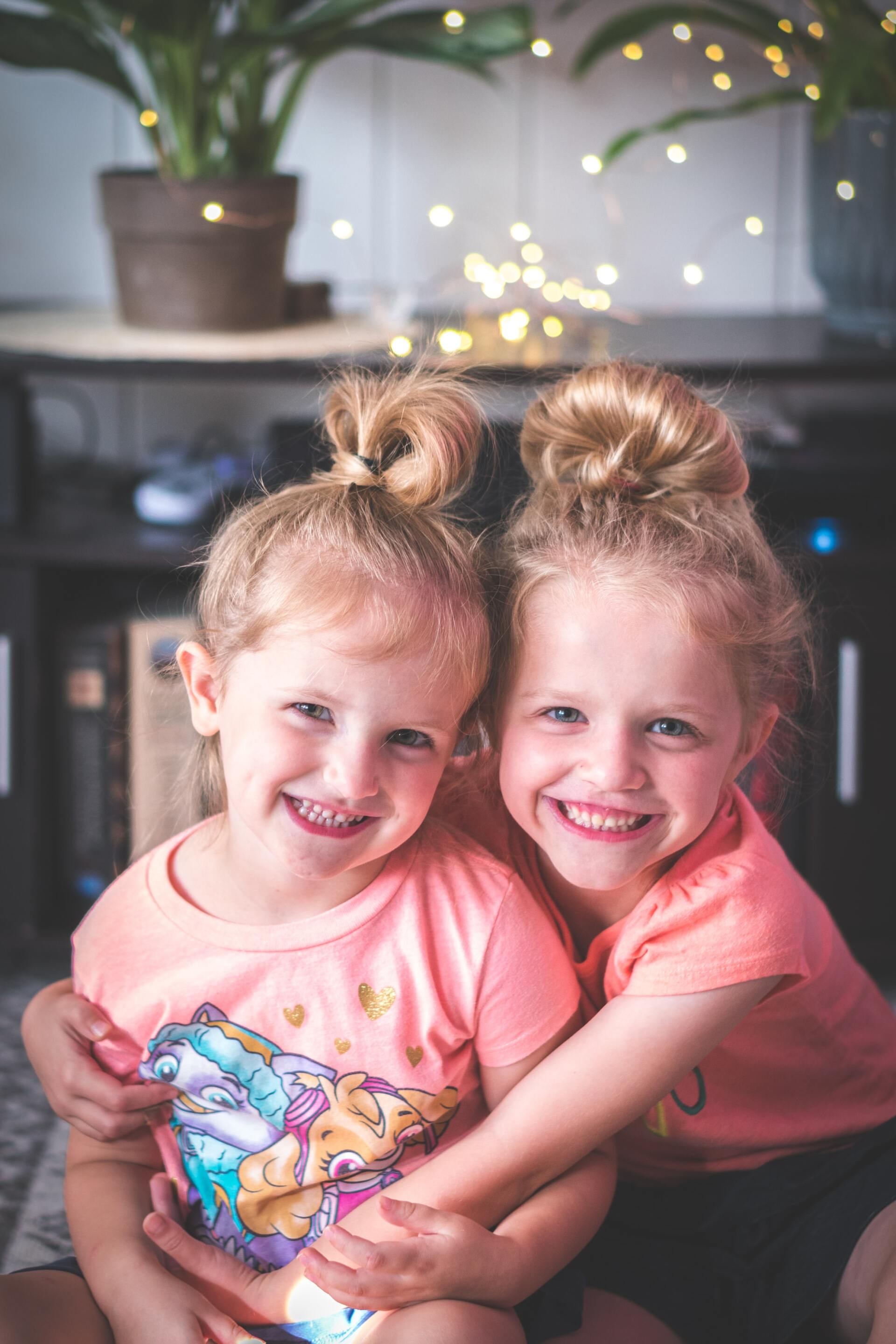 Two young girls with blonde buns smile. They wear pink shirts, sitting on a rug with fairy lights in the background.