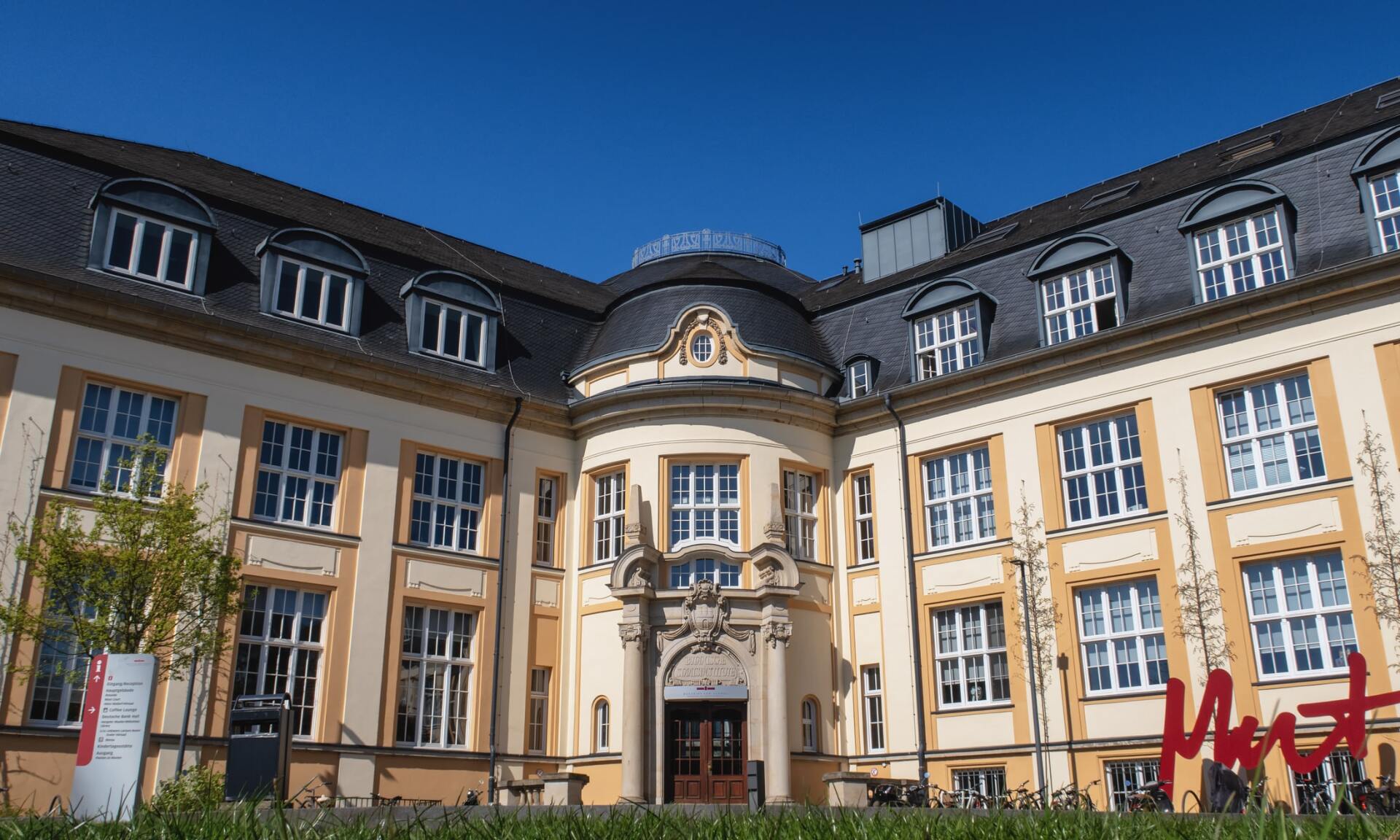 Beige building with many windows, central rounded entrance, dark roof, blue sky.