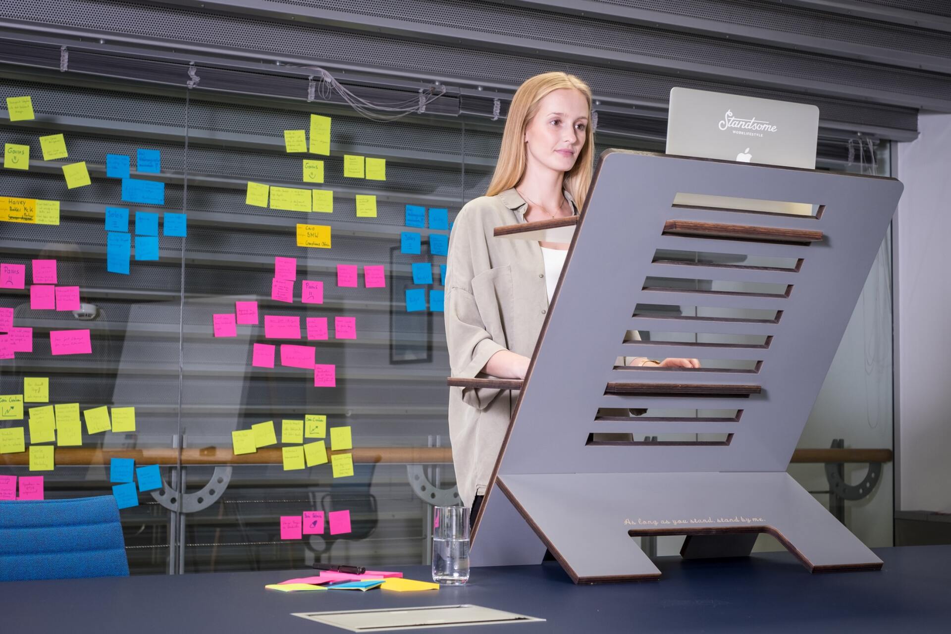 A woman is standing at a standing desk in front of a wall with sticky notes.