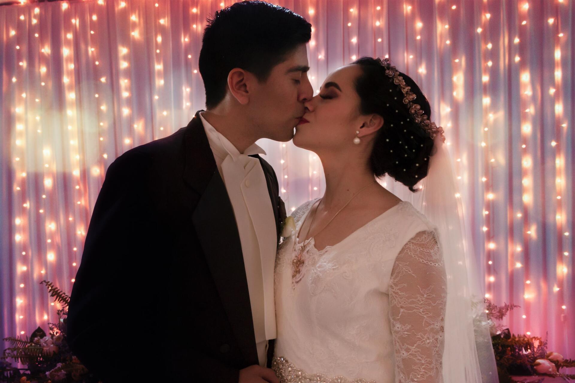 Una novia y un novio besándose delante de una cortina de luces.