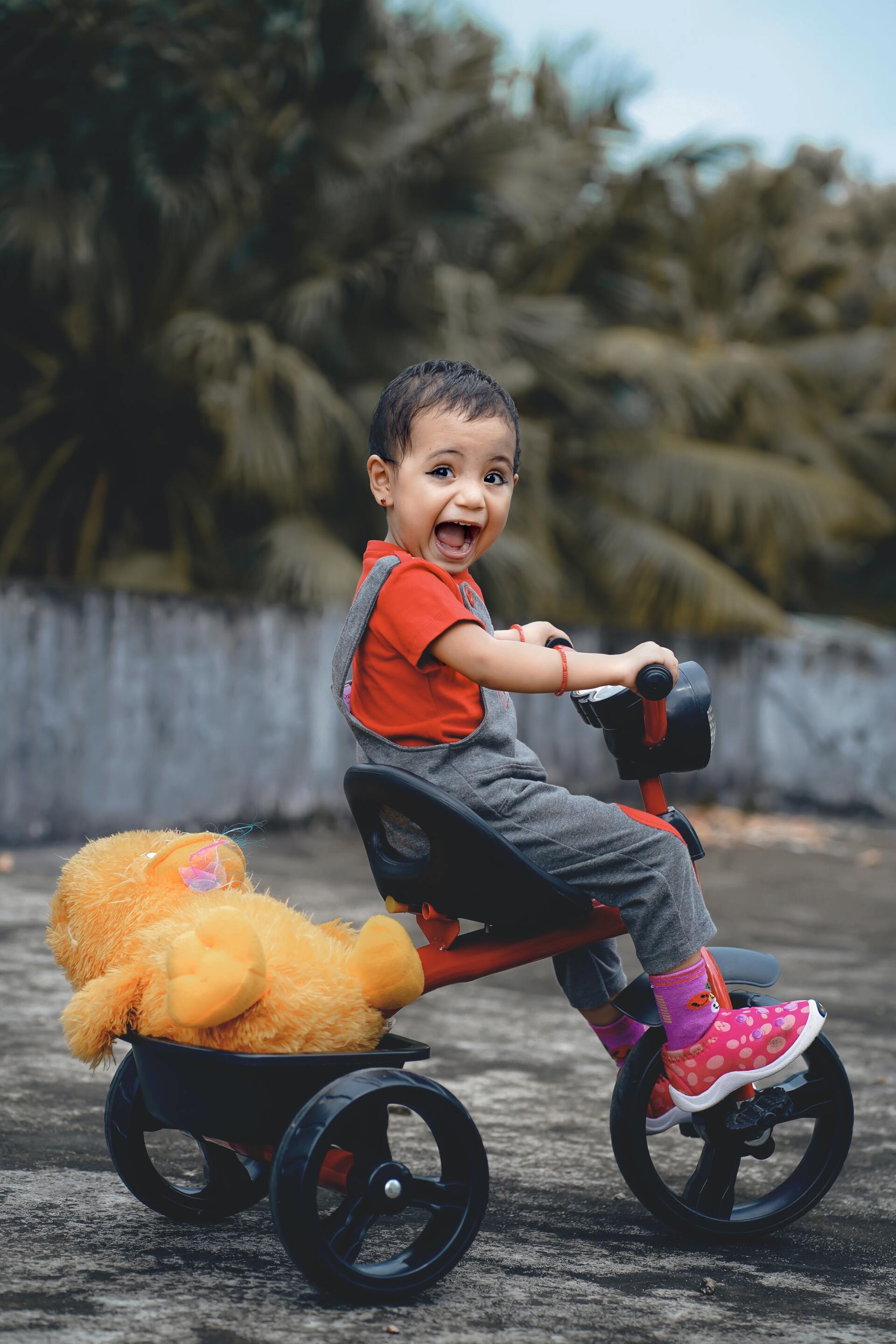 Child on a tricycle, smiling widely, with a teddy bear in the back. Wearing red shirt and rain boots. Outdoors.