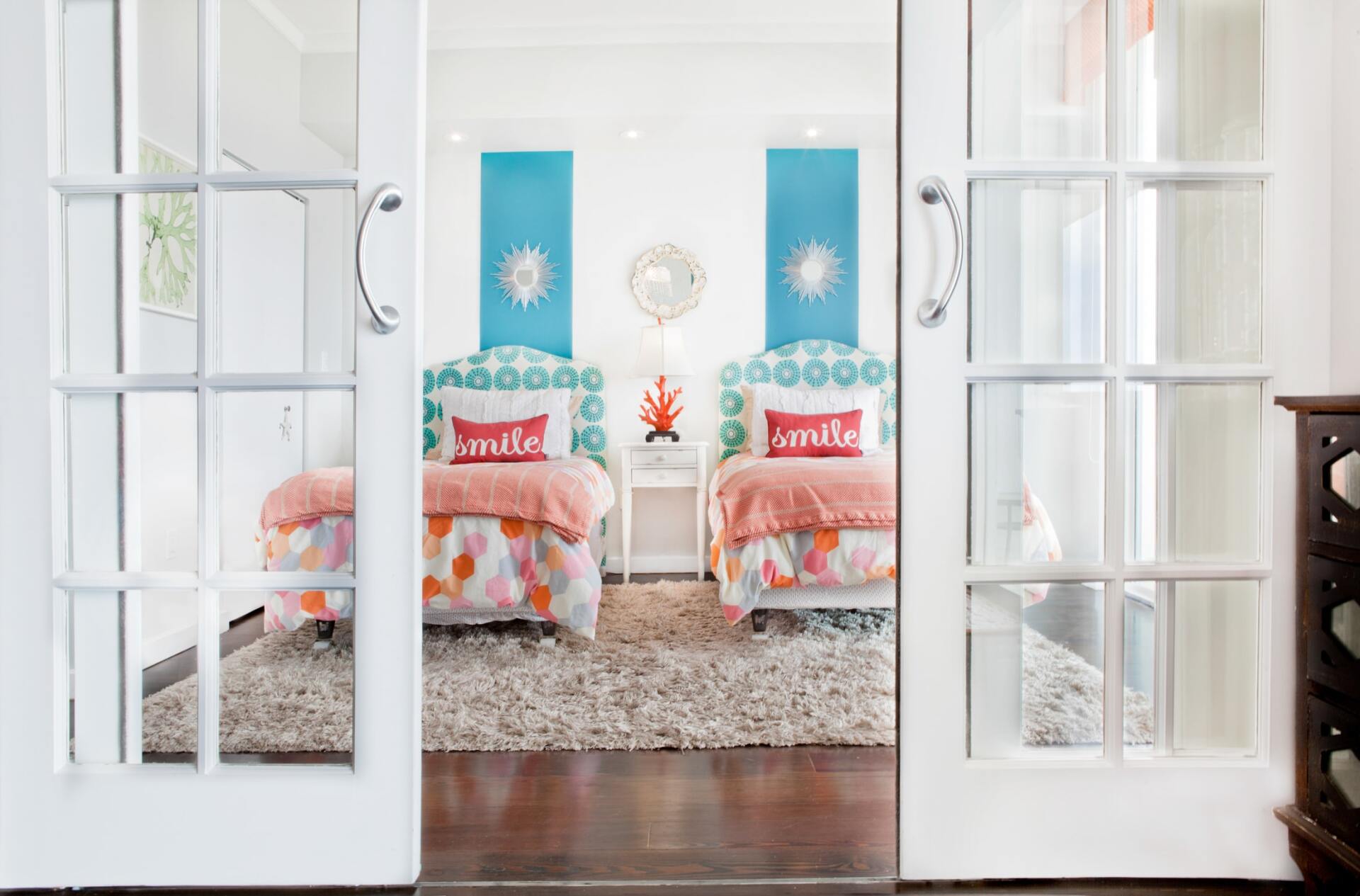 Two twin beds in a white room, viewed through open French doors. Each bed has a patterned headboard and orange bedding.