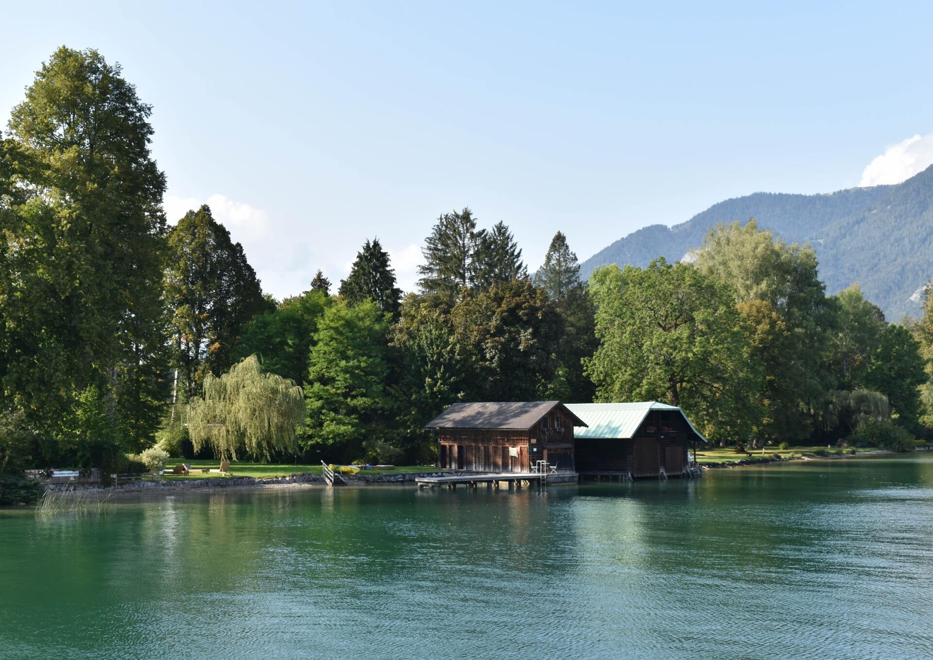 Bootshaus am Seeufer, neben ruhigem Wasser, umgeben von grünen Bäumen und Bergen unter klarem Himmel
