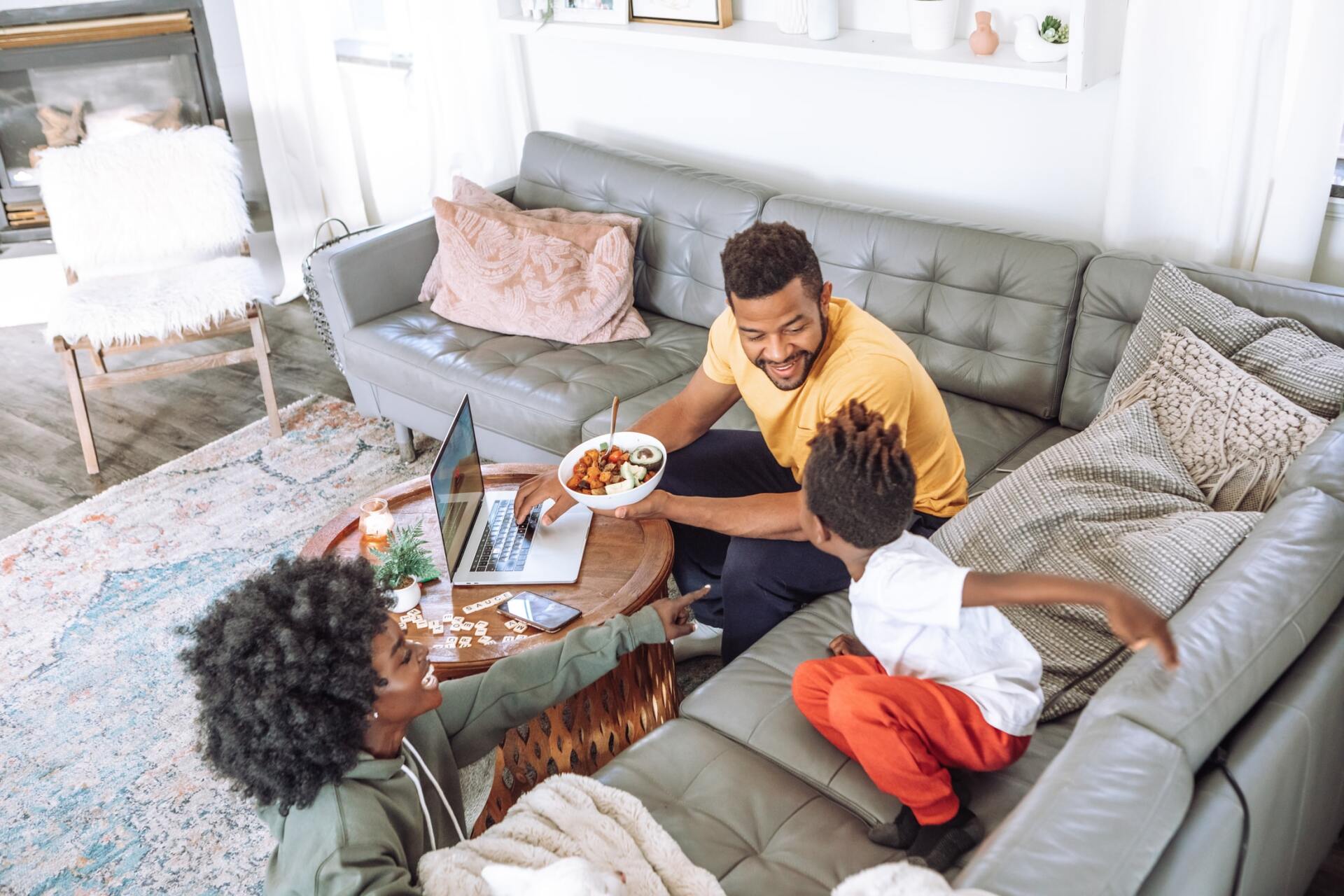 A family is sitting on a couch eating food and using a laptop.