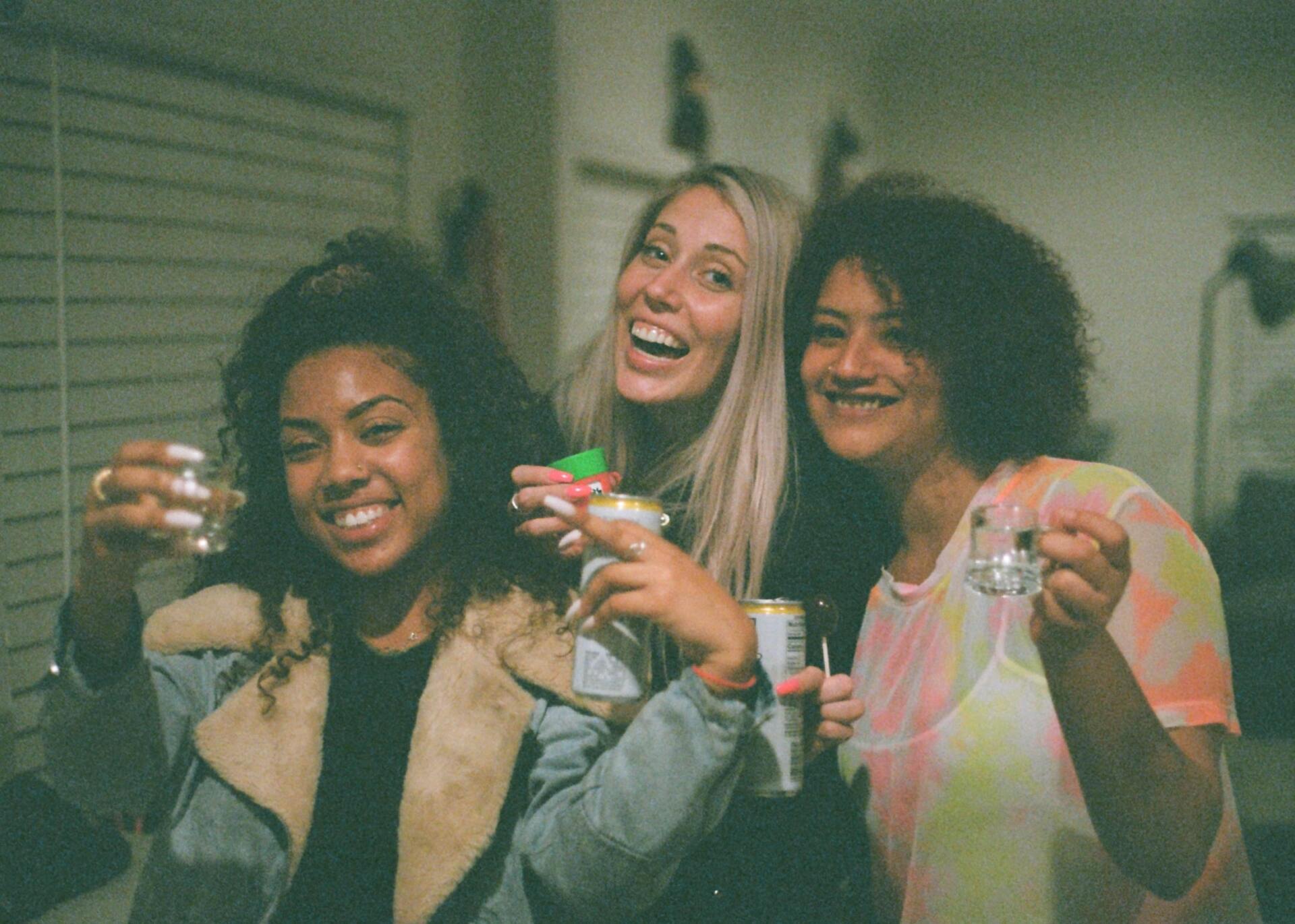 Three friends smile and hold up small glasses for a toast in a brightly lit indoor space.