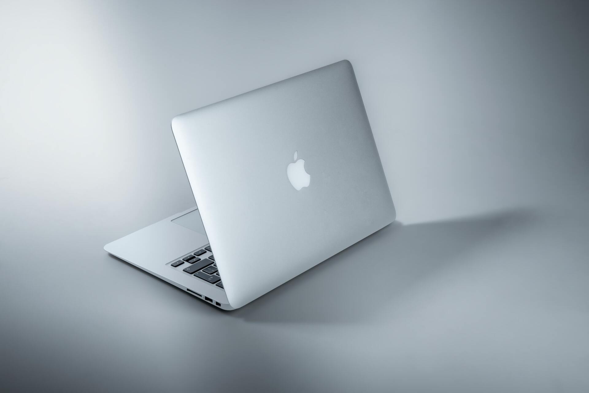 Zilverkleurige laptop met oplichtend Apple-logo op een grijze achtergrond, gedeeltelijk open en van de zijkant gezien.
