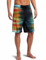 Man wearing rainbow-striped board shorts standing barefoot on a white background