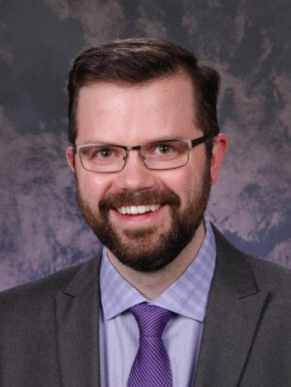 Portrait of a smiling man in a gray suit, purple tie, and glasses against a studio backdrop