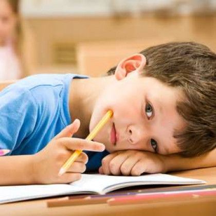 Boy in a blue shirt resting his head on an open notebook, holding a pencil at a desk.