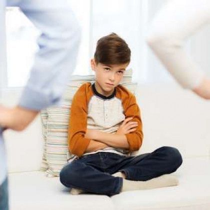 Child sitting cross-legged with arms crossed, looking upset, in a bright room with blurred adults nearby
