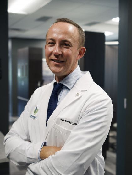 Doctor in white coat standing with arms crossed in a hospital hallway, smiling.