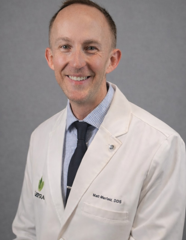 Doctor in white coat standing with arms crossed in a hospital hallway, smiling.