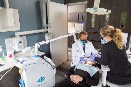 Dentist treating a patient in a modern clinic, with an assistant beside him and a machine nearby.