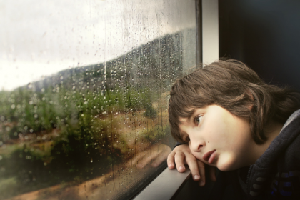 Child leaning on a train window, gazing at rain-soaked scenery outside