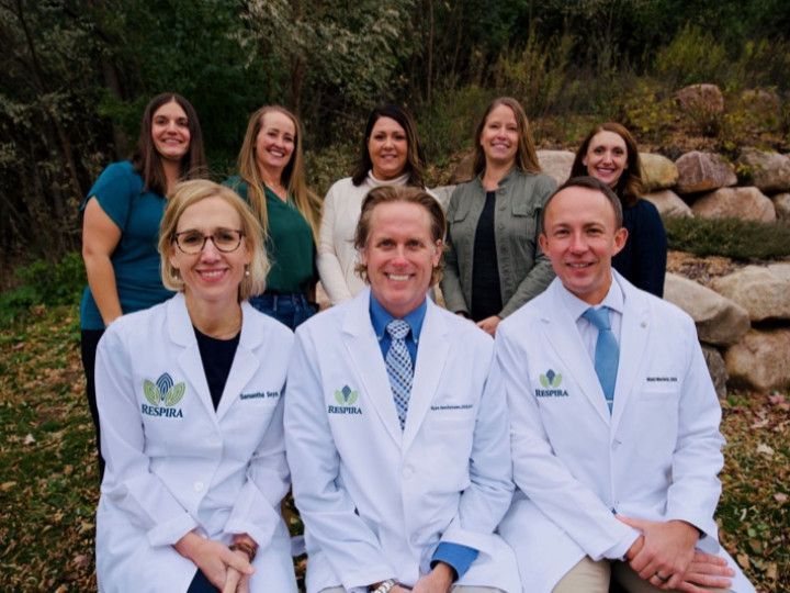 Group of six medical professionals in white coats posing outdoors in front of greenery.