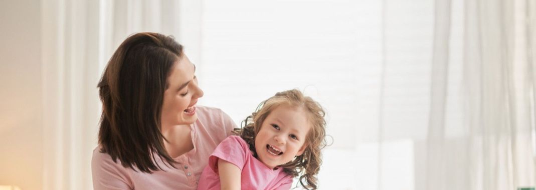 Woman in pink holding a smiling child on a bright couch by a sheer-curtained window