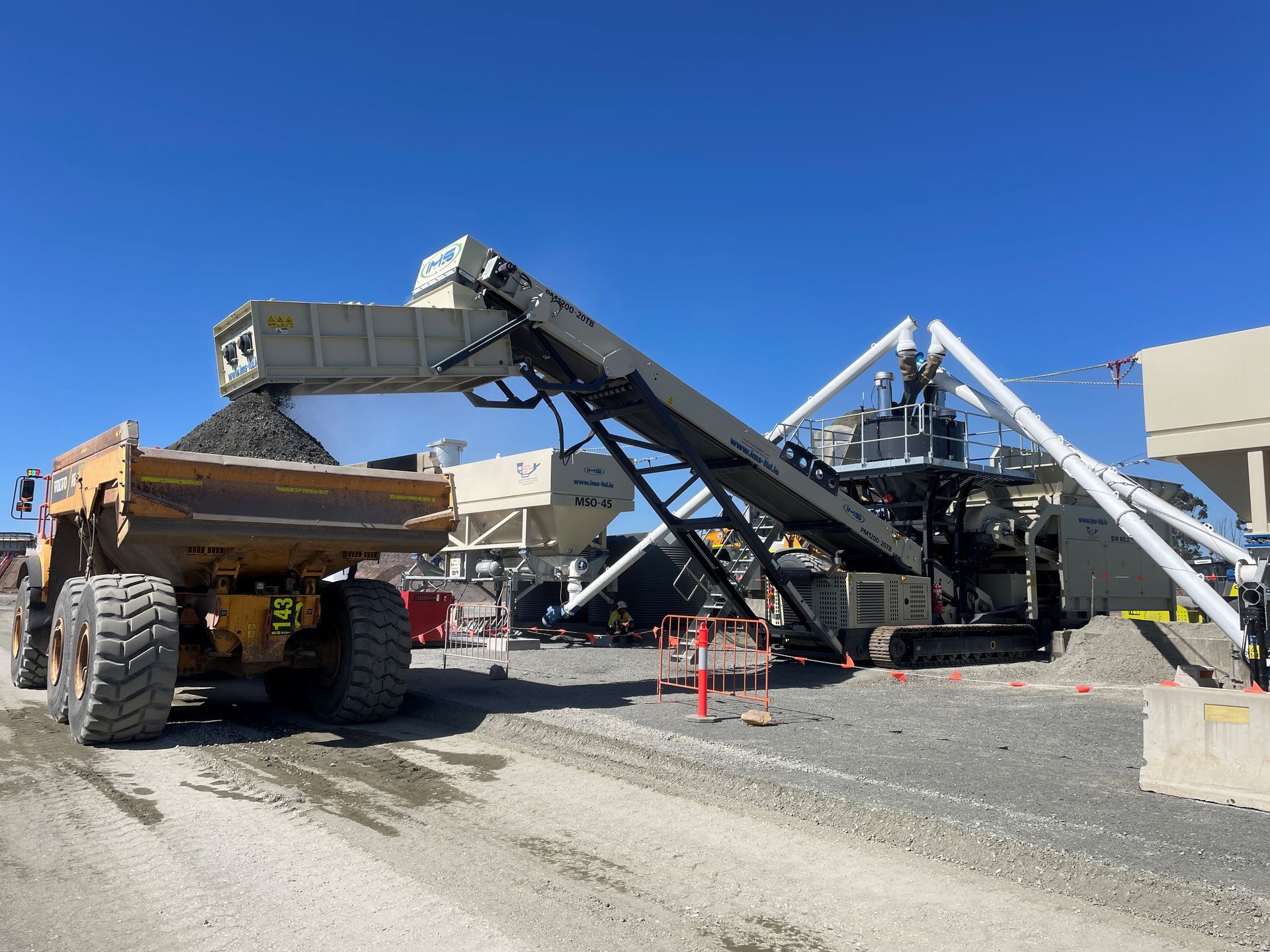 A yellow dump truck is being loaded with dark material from a conveyor belt at a construction site, sunny day.