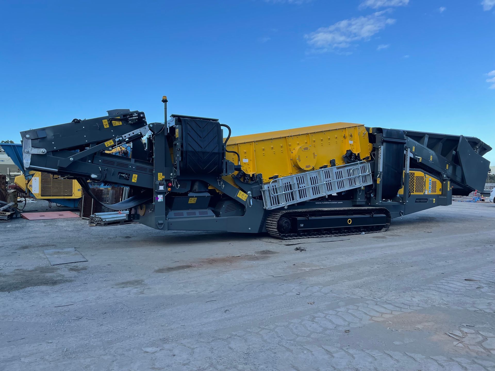 A large yellow and gray mobile rock crusher on tracks, parked on a light-colored surface against a blue sky.