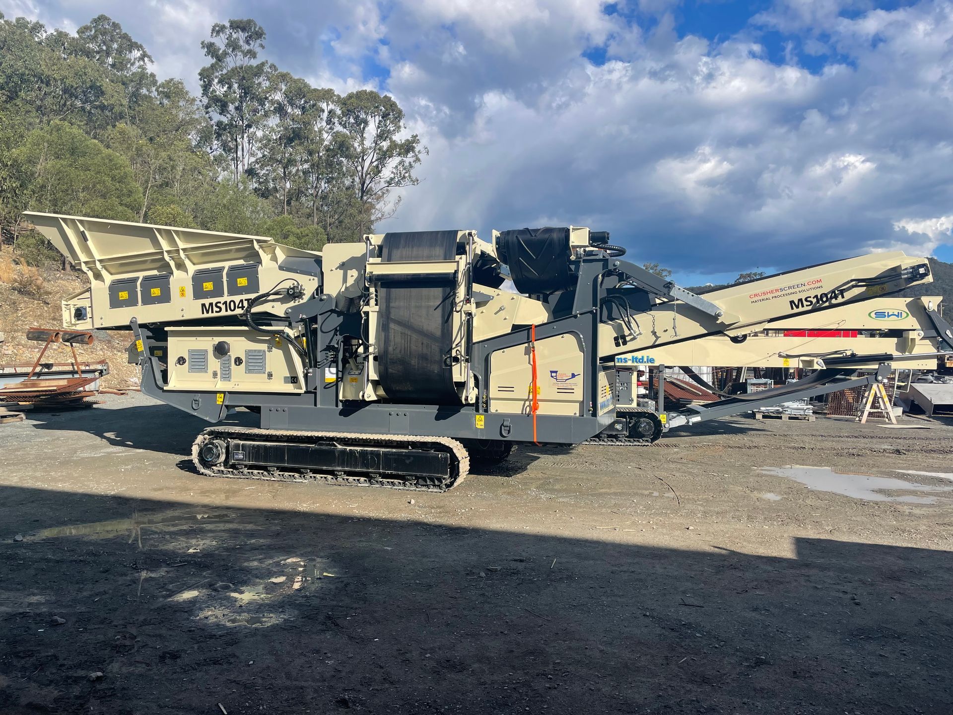 A large, beige and gray mobile rock crusher on a gravel surface, likely at a quarry or construction site, with trees in the background.