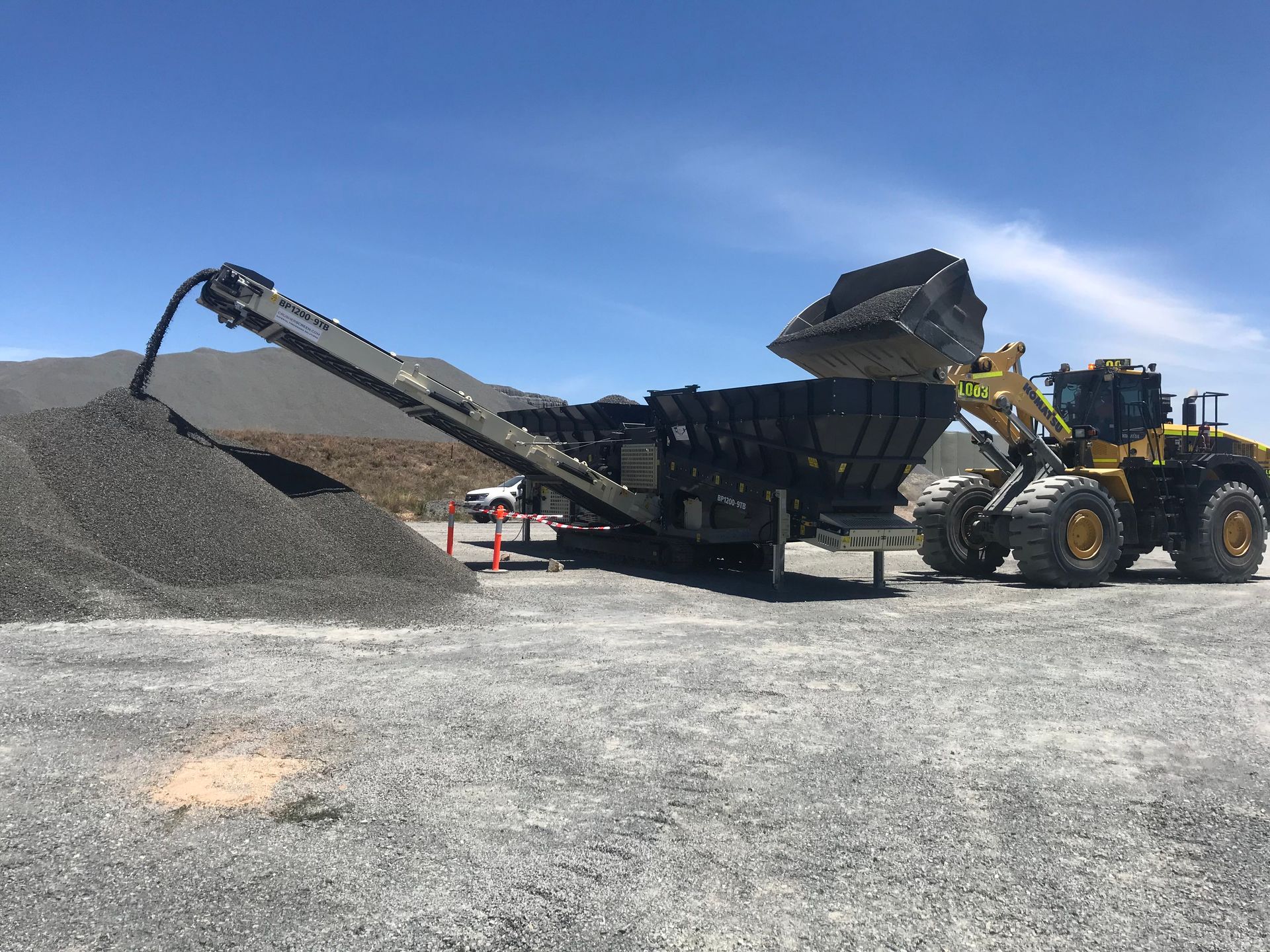 A yellow front-end loader deposits rocks into a rock screening machine, creating a pile of gravel against a blue sky.