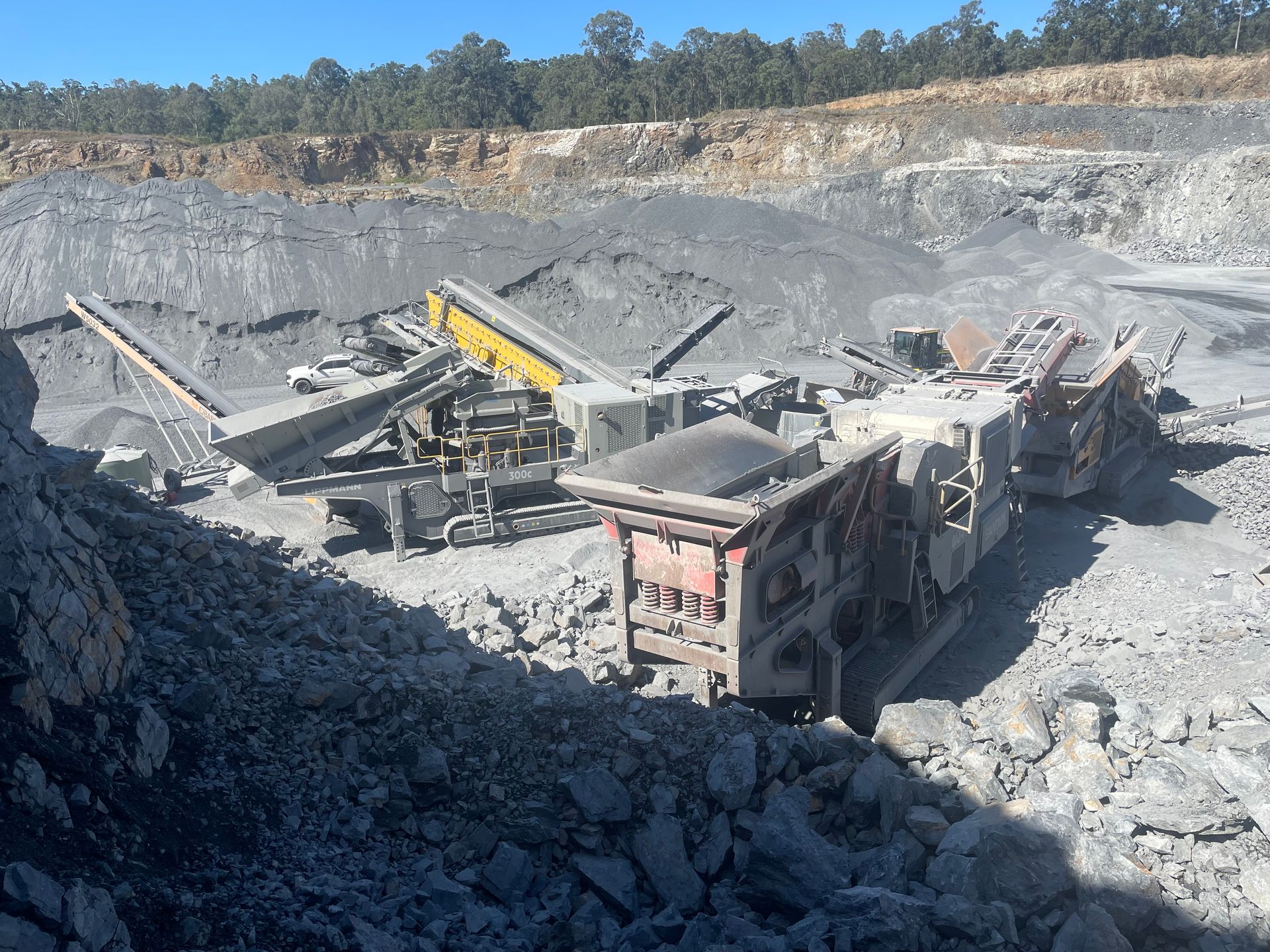 Quarry machinery processing rocks at an outdoor site. Grey and yellow equipment. Bright sunlight.