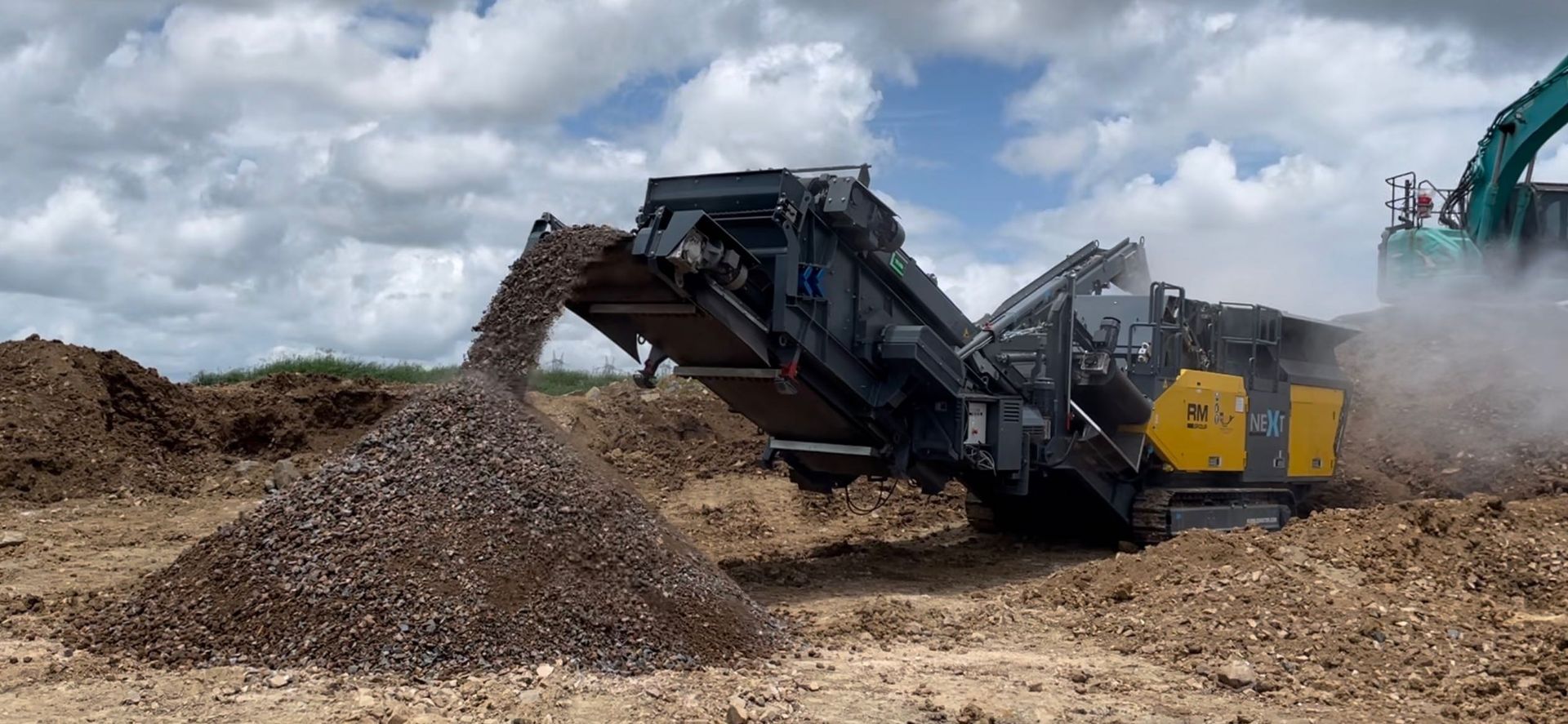 A large machine crushing rocks, dumping a pile of gravel on a brown, arid landscape under a cloudy sky.