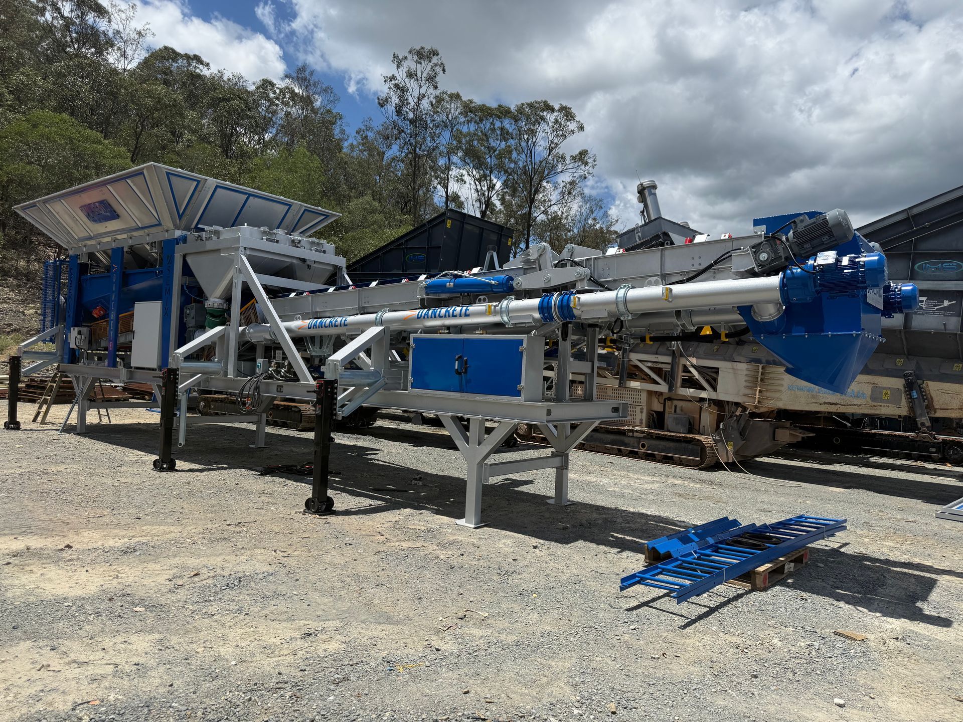 Blue and white industrial machine, outdoors on gravel. Sky and trees in the background.