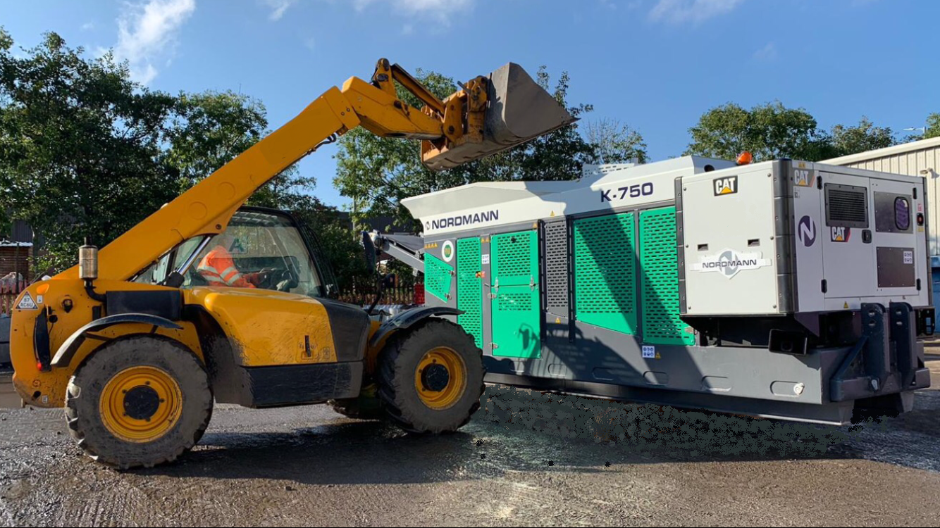 A yellow telehandler loads material into a green and white industrial machine outdoors on a sunny day.