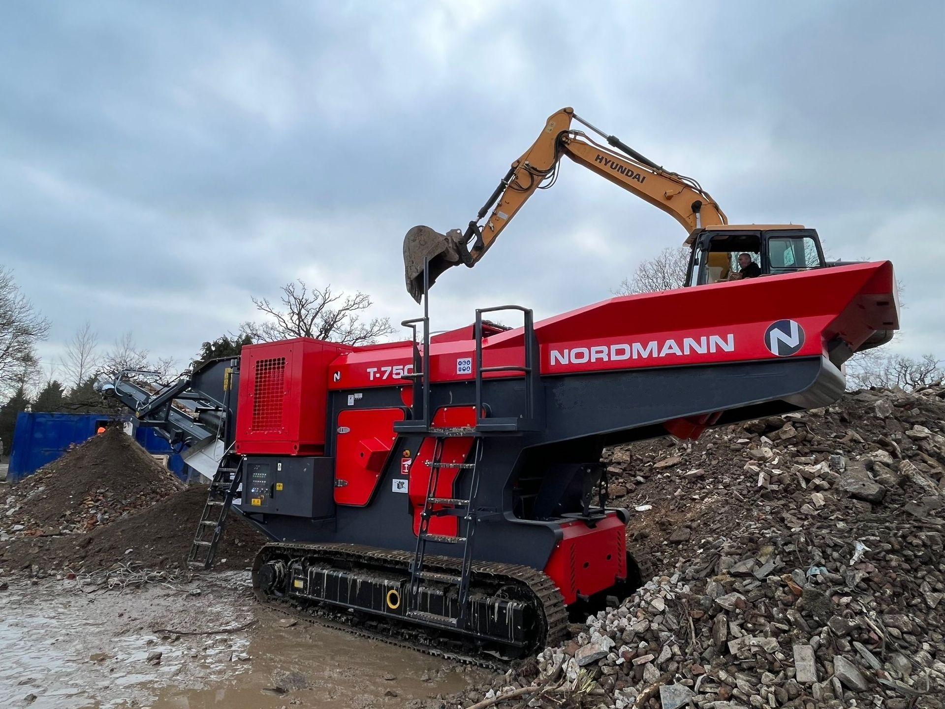 A red Nordmann T-7212 mobile shredder is loaded by an excavator on a debris pile.