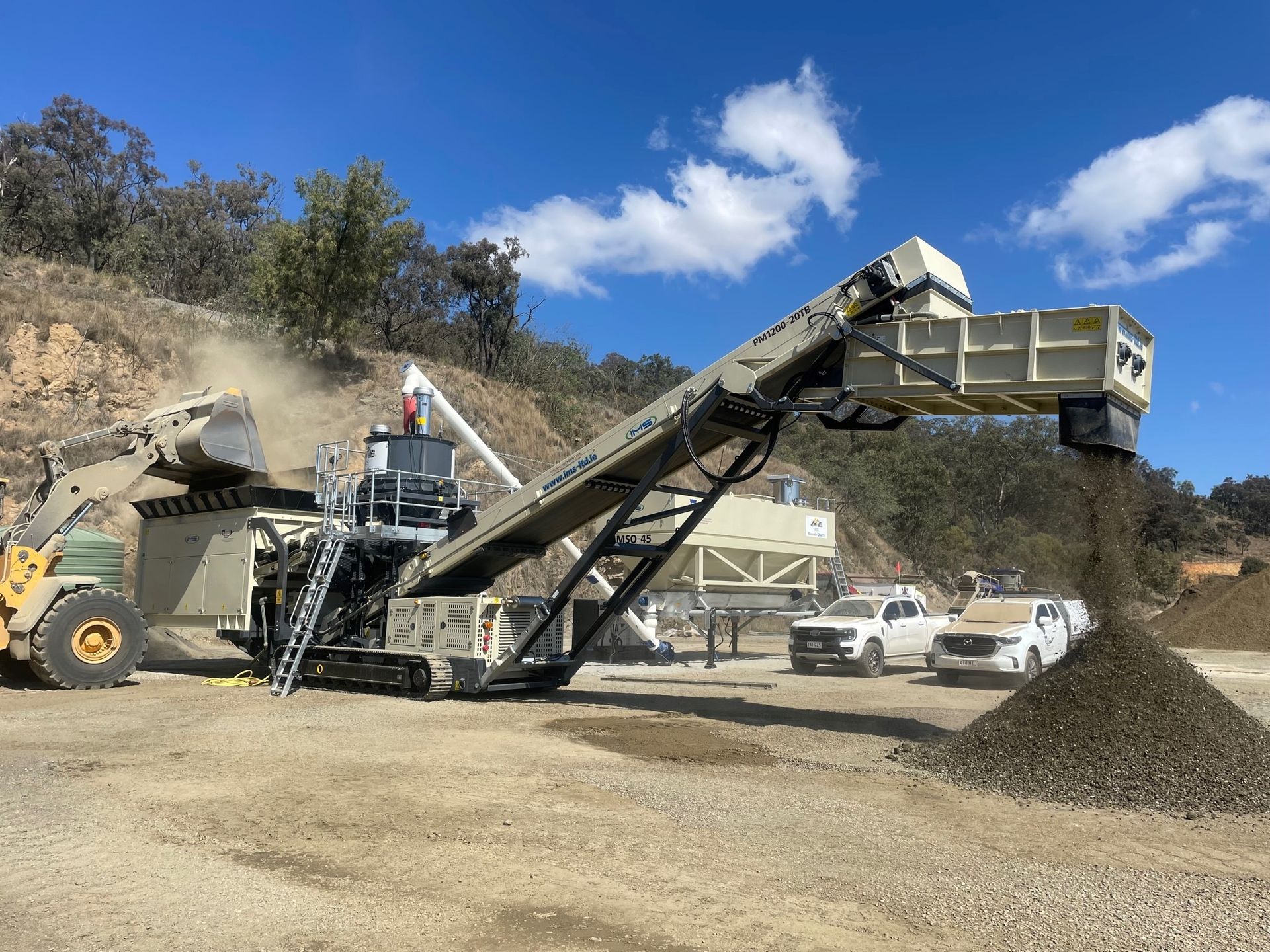 A large, mobile rock crushing machine in operation at a quarry, with a pile of crushed stone and white vehicles parked nearby.
