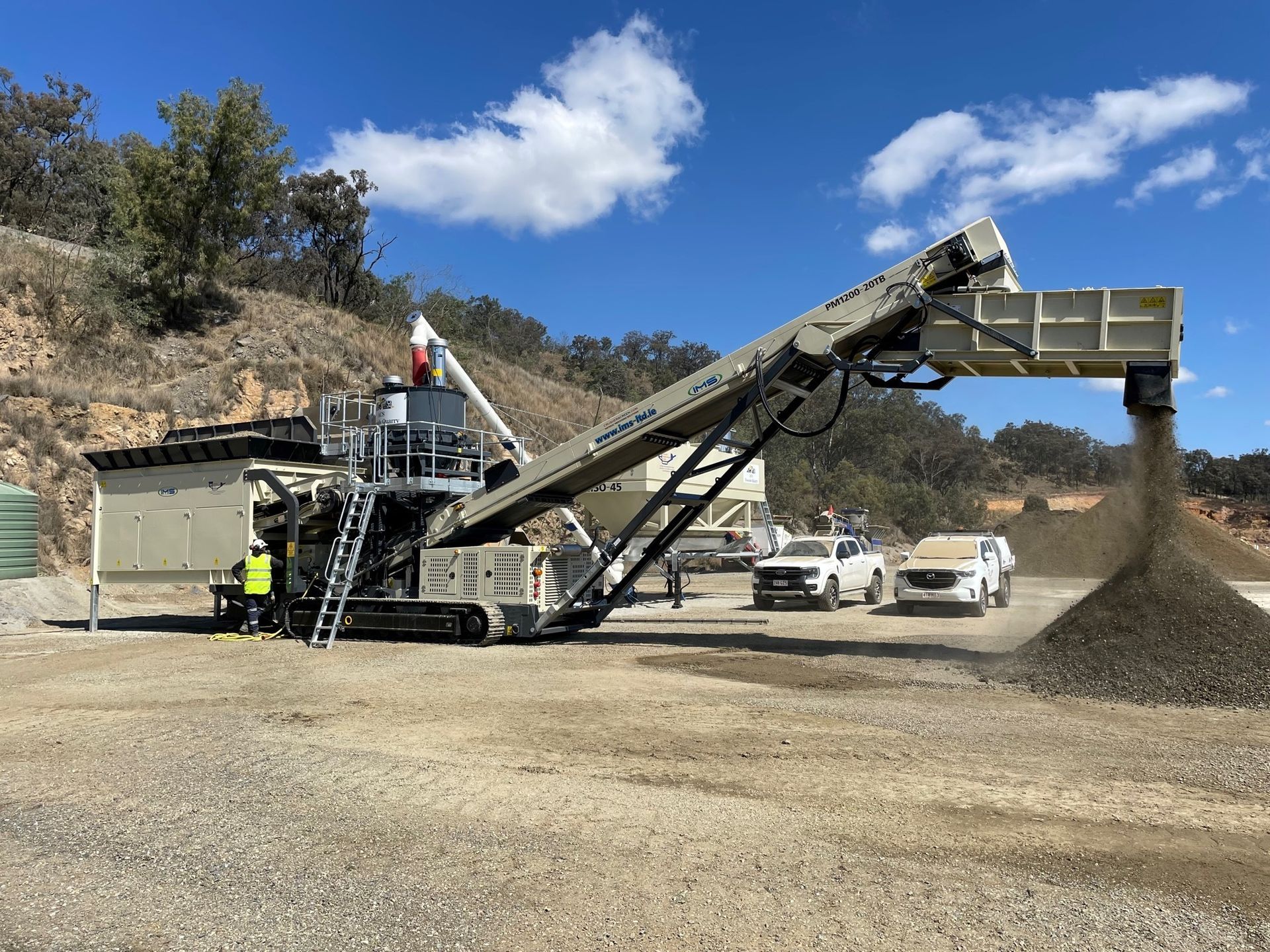 A large mobile rock crusher sorts gravel into a pile outdoors on a sunny day. Two white trucks are parked nearby.