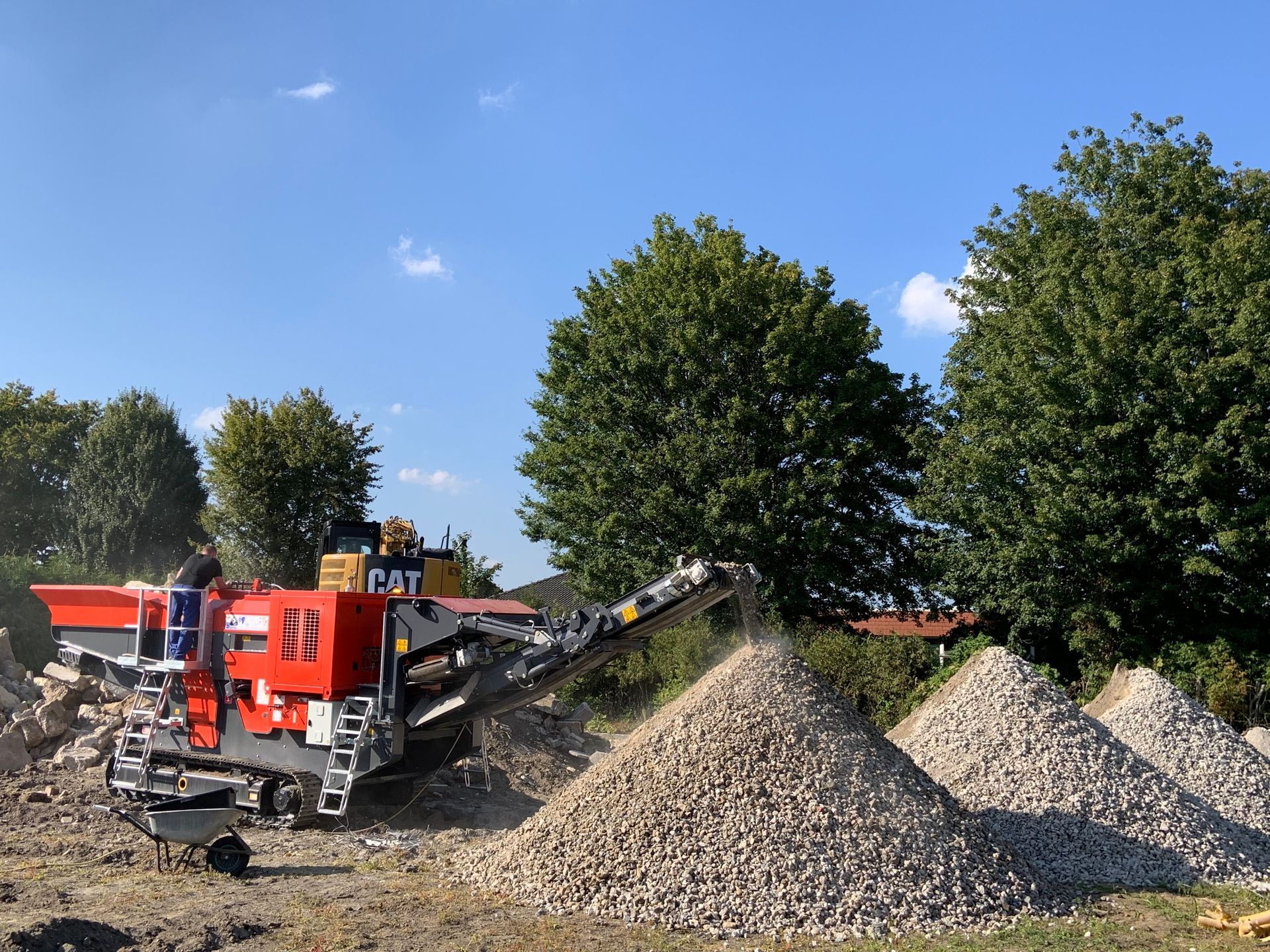 A red industrial rock crusher working in an outdoor setting, with piles of crushed rock and trees in the background.