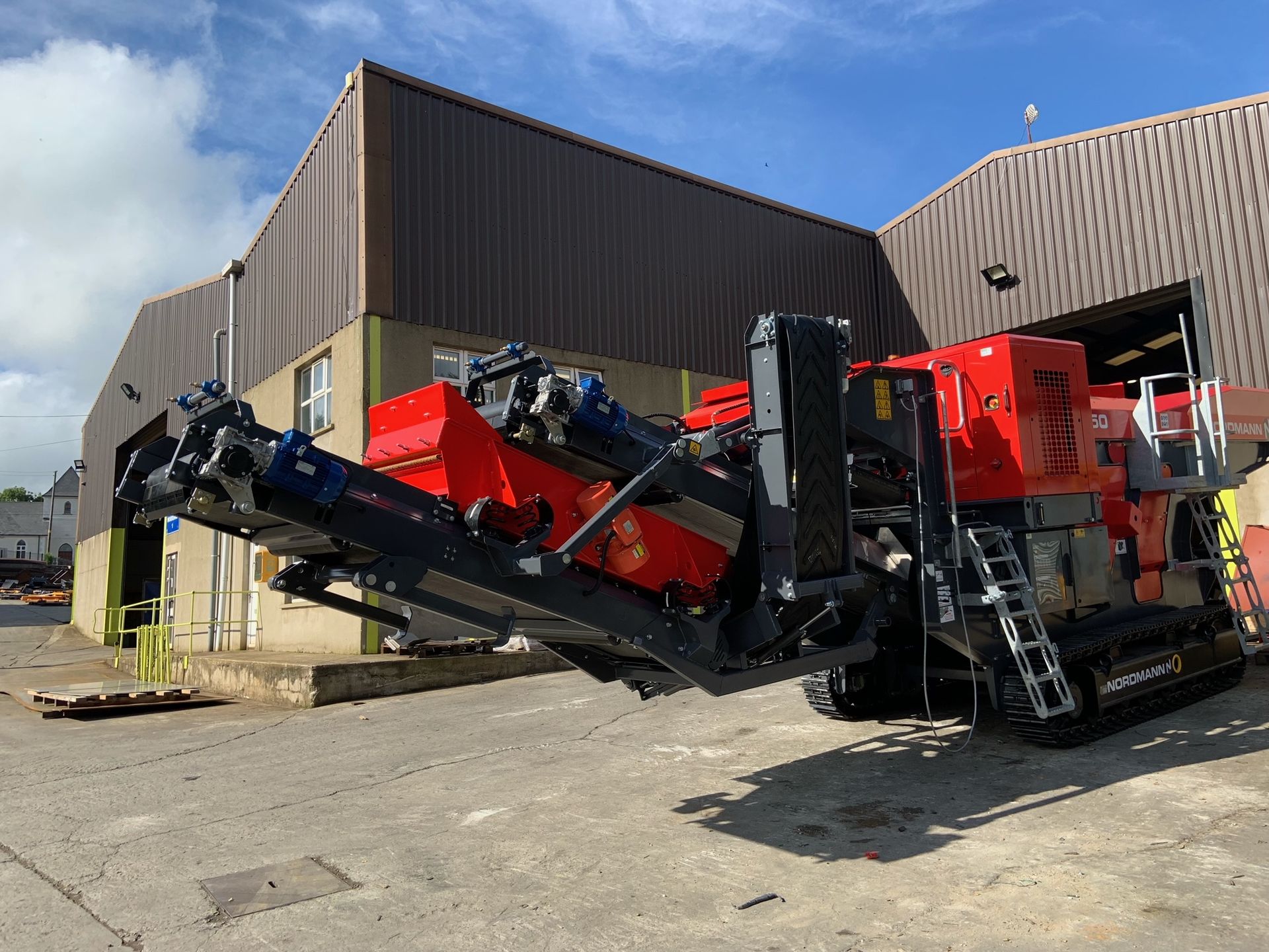 Red and black industrial machine on treads outside a building on a sunny day.